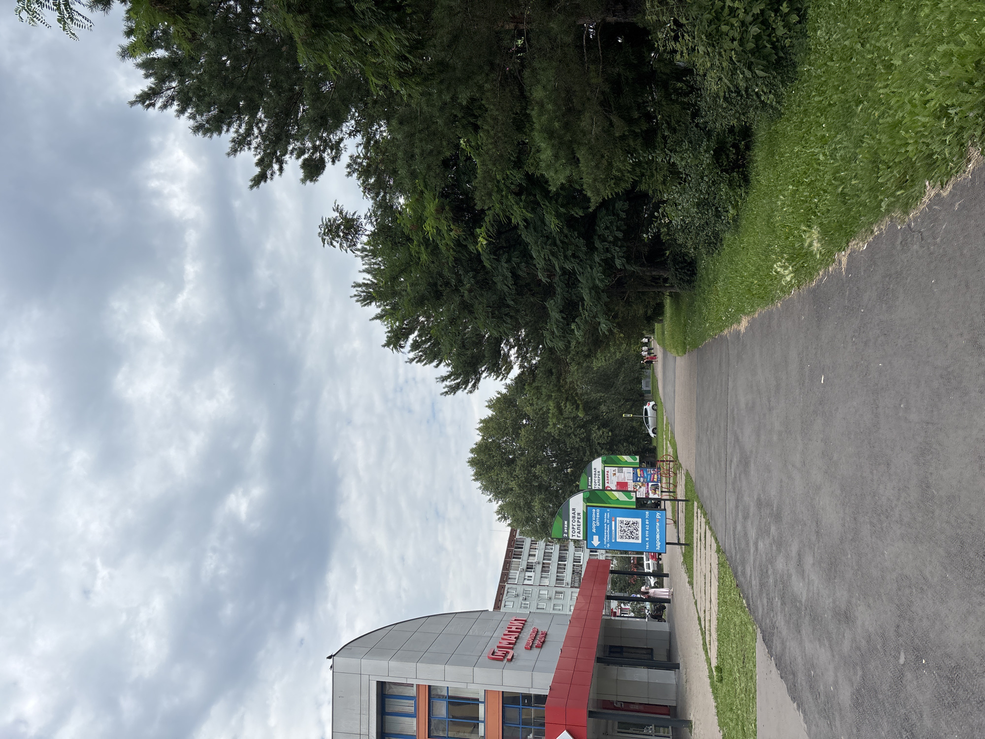 An outdoor, eye-level, medium shot captures a paved pedestrian path leading into the distance, bordered on the left by a building and on the right by a dense line of trees and green foliage. The sky above is overcast with heavy, gray clouds.
On the left, a modern building with a curved facade dominates the foreground. It features a red and white color scheme, with large windows on the upper floors and a red awning over the ground-level entrance. A red "M" logo, likely for a store, is visible on the building. Beside the building, a blue sign displays a QR code and Russian text, possibly indicating directions or information. Several parked cars are visible on the street adjacent to the building.
On the right side of the path, a variety of trees, including tall evergreens and leafy deciduous trees, create a wall of greenery. The ground beneath the trees is covered in lush grass and smaller bushes.
In the distance, more buildings and trees can be seen, suggesting an urban or suburban environment. A few people are walking on the path in the distance, some in casual attire. The overall atmosphere is subdued, likely due to the cloudy weather.
The signs in the image contain Russian text, indicating the location is likely in Russia. One sign specifically mentions "г. Набережные Челны" which translates to "Naberezhnye Chelny," a city in Russia. The text on the blue sign mentions "Аптека" (pharmacy) and "Здрависти.ру," which is a Russian online pharmacy. Another sign indicates "Торговая Галерея" (Shopping Gallery). These details confirm the location as Naberezhnye Chelny, Russia, specifically near a shopping gallery and an apartment building.