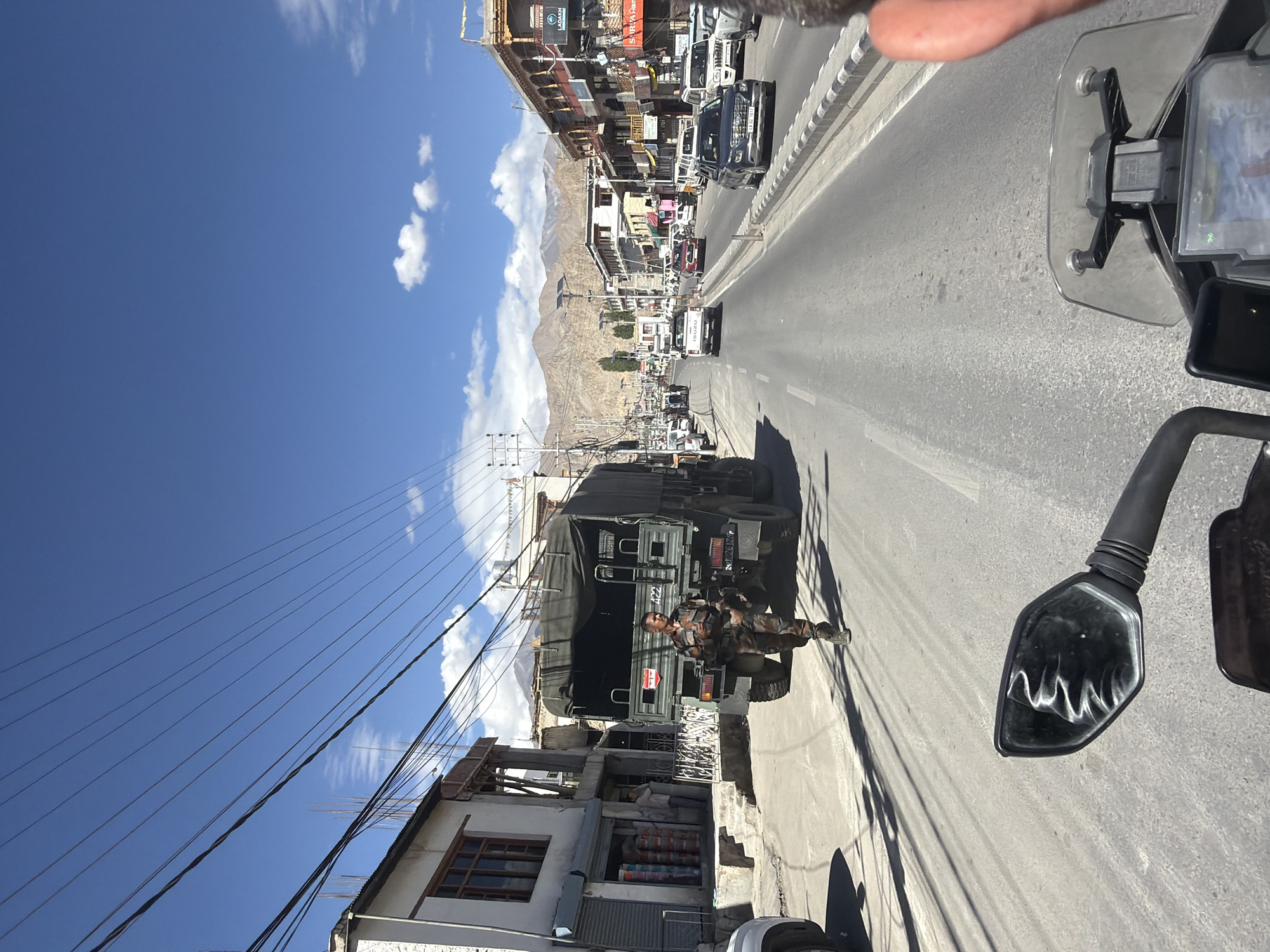 The image depicts a street scene in Leh, India, on a clear day with scattered clouds. The foreground shows the handlebars and a partial view of a motorcycle's windshield and display. A soldier in camouflage uniform is walking on the right side of the road, in front of a large military truck. The truck has a canvas cover and the number "422" visible on its rear. Various cars and other vehicles are present on the road, with buildings lining both sides. The architecture of the buildings suggests a regional style, with some featuring wooden accents. In the background, mountains are visible, characteristic of the Himalayan region. The road surface is asphalt, and utility poles with numerous wires are prominent throughout the scene. The overall atmosphere appears to be one of everyday activity in a town with a significant military presence.