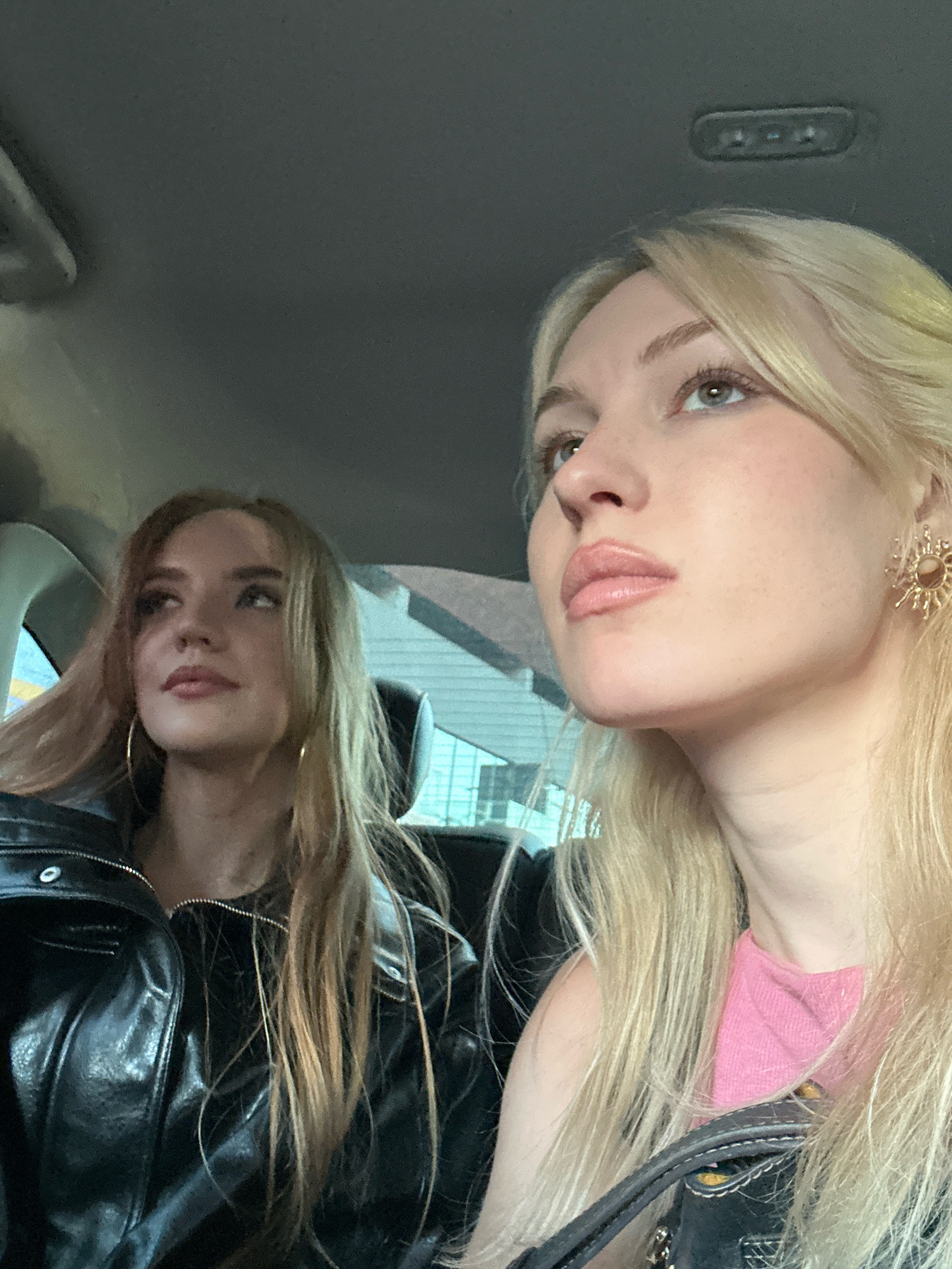 Two young women are seated in the back of a vehicle, likely a car, looking upwards and to their right. The woman in the foreground, with blonde hair and a pink top, is in profile, her gaze directed away from the camera. She is wearing a sun-shaped earring on her right ear. The woman behind her, with lighter brown hair and wearing a black leather jacket, is also looking upwards and to her right. The interior of the vehicle appears to be a standard car interior with a dark-colored headliner. The background suggests an urban environment, possibly with buildings visible through the car windows, hinting at a location within the city of Tbilisi, Georgia. The lighting suggests it is daytime, possibly overcast.