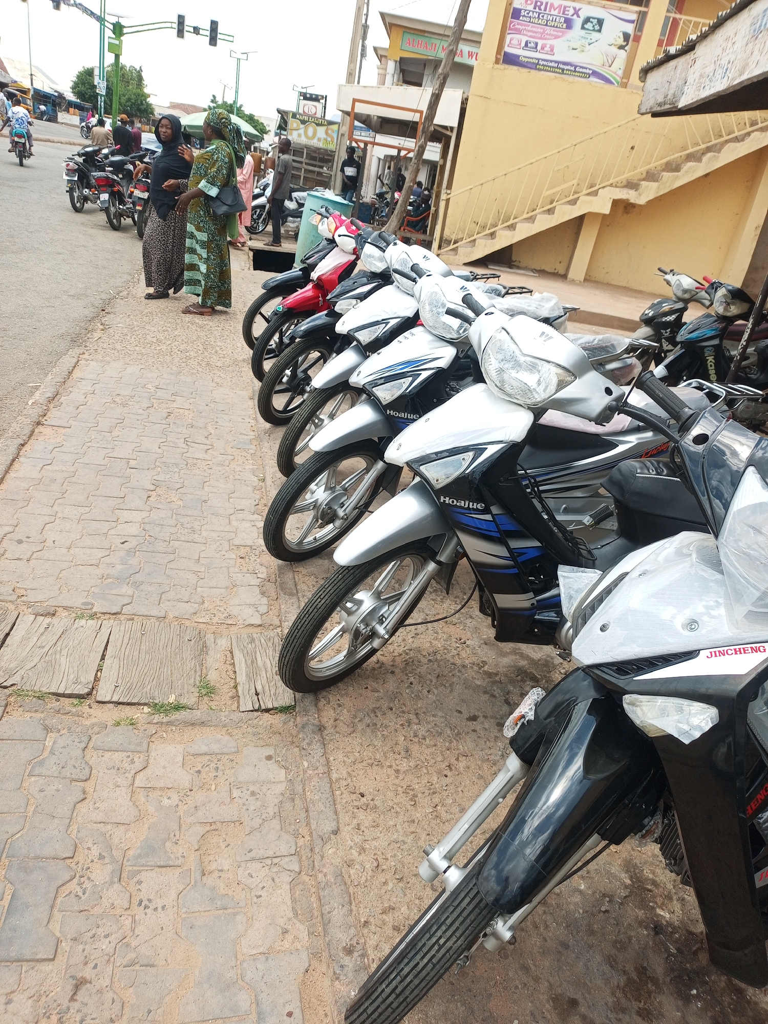 The image shows a street scene in the city of Arawa, Nigeria, with a focus on a row of parked motorcycles. Two women are standing on the sidewalk on the left side of the image, facing away from the camera. The woman on the left is wearing a black hijab and a patterned skirt, while the woman on the right is wearing a green and patterned dress and a headscarf. Several motorcycles are parked in a line on the sidewalk. The motorcycles are varied in color, with several silver and black models prominently featured. In the background, there are buildings and a street with a few people and motorcycles visible. The environment appears to be a busy commercial or residential area.