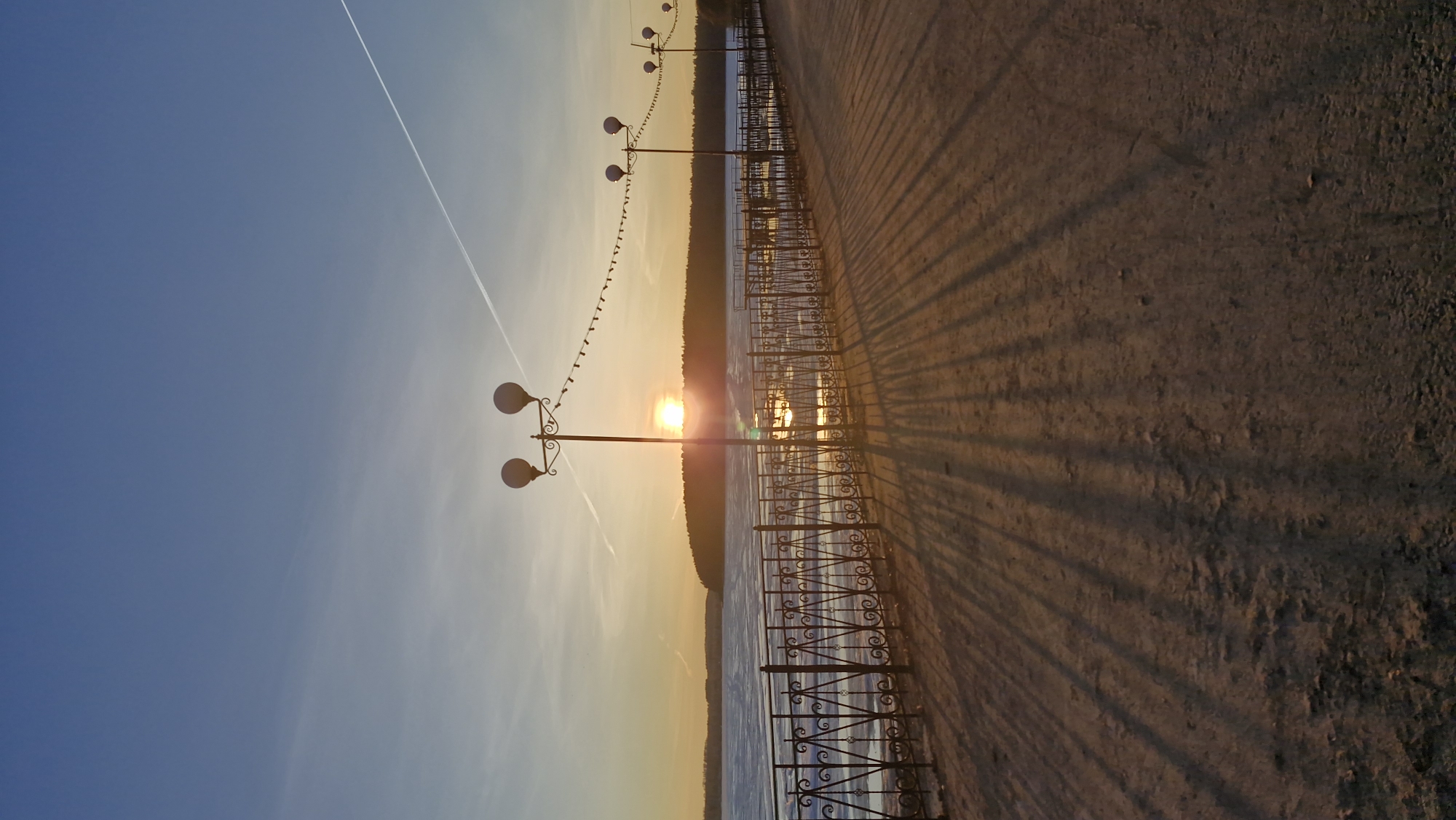A landscape photograph of a promenade in Otchor, Russia, at sunset. The sky is blue near the top left, fading to a yellow and orange gradient above the horizon. A contrail bisects the sky diagonally. The sun is setting behind a line of trees in the distance, its reflection visible in the water. Along the promenade, there are ornate black metal railings and evenly spaced lampposts with spherical lamps. Small decorative flags are strung between the posts. The ground on the right side of the image is an earthy tone, cast with long, linear shadows from the railings and posts.