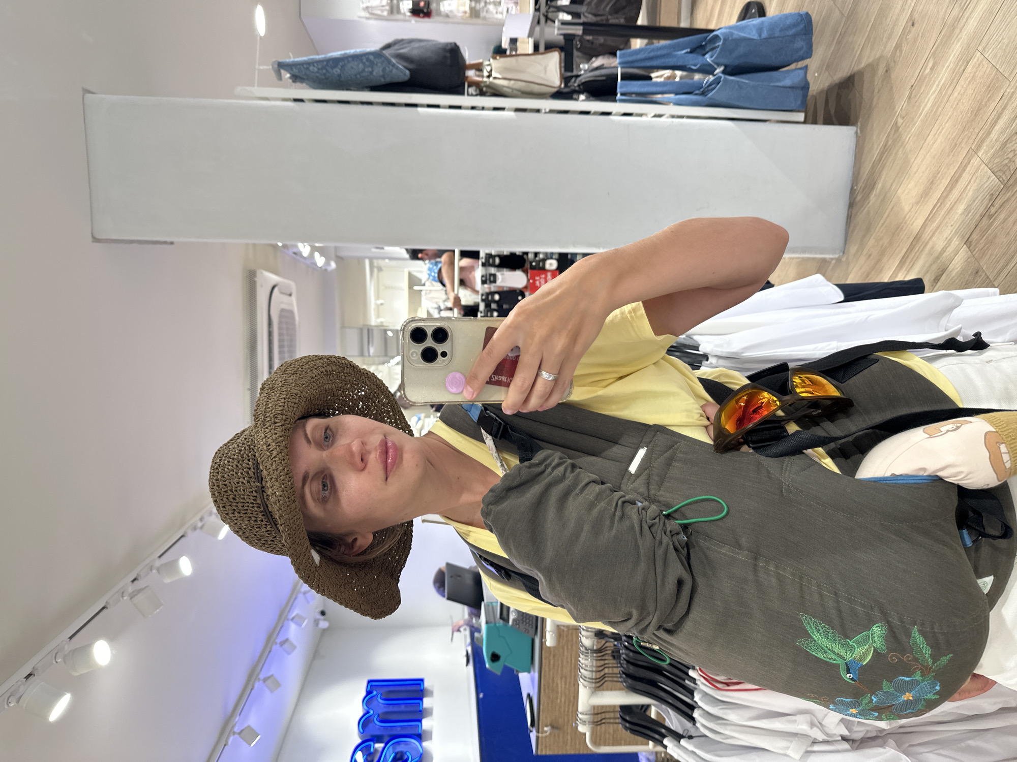 A woman in a hat and yellow shirt takes a selfie in a clothing store in Fethiye, Turkey. She holds an iPhone with a pink object on the back. She is wearing a baby carrier with a hummingbird design. Sunglasses rest on top of the carrier. In the background are racks of clothing and store fixtures. Overhead are track lights.