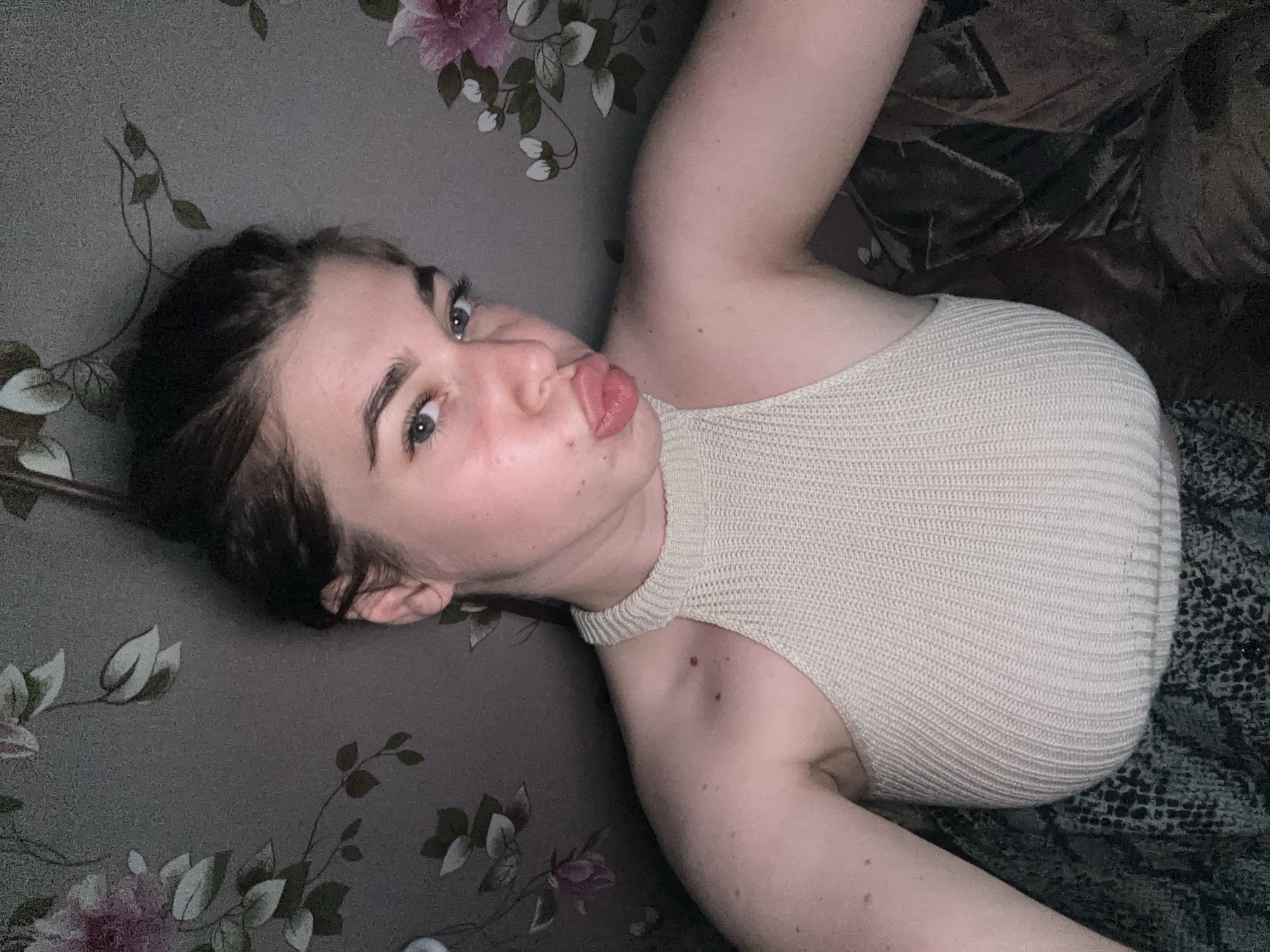 The image shows a young woman with her hair in a bun, pouting her lips. She is wearing a cream-colored, ribbed halter top and a patterned skirt. She is positioned in front of a wall with floral wallpaper. The background is dark and dimly lit, with a faint floral pattern visible. There is a brown, possibly velvet, fabric to the right of the woman. The scene appears to be indoors, likely in a domestic setting. There is no indication of the location being in the city of Мещёры, Russia.