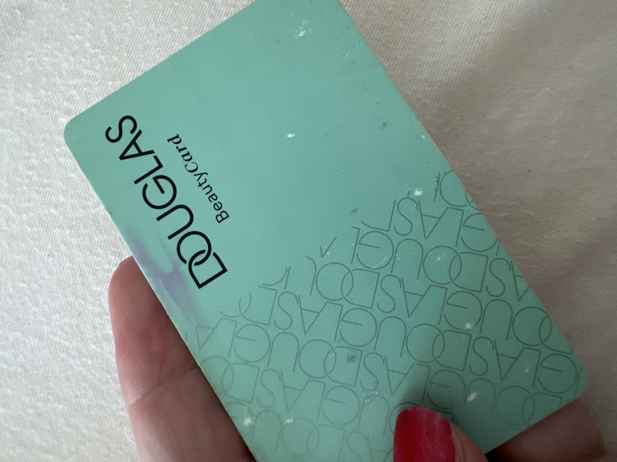 An image shows a close-up of a hand holding a turquoise Douglas Beauty Card. The card has the Douglas logo, which is two interlocking 'D' shapes, printed in black, and the text "Beauty Card" below it. The background of the card features a repeating pattern of the word "Douglas" in a lighter shade of turquoise. The hand holding the card has a red fingernail polish visible on one finger. The card is lying on a textured, light-colored surface, likely a tablecloth or fabric. There are a few small white specks on the surface of the card, possibly dust or smudges. The image does not provide any information about the location in Krefeld, Germany.