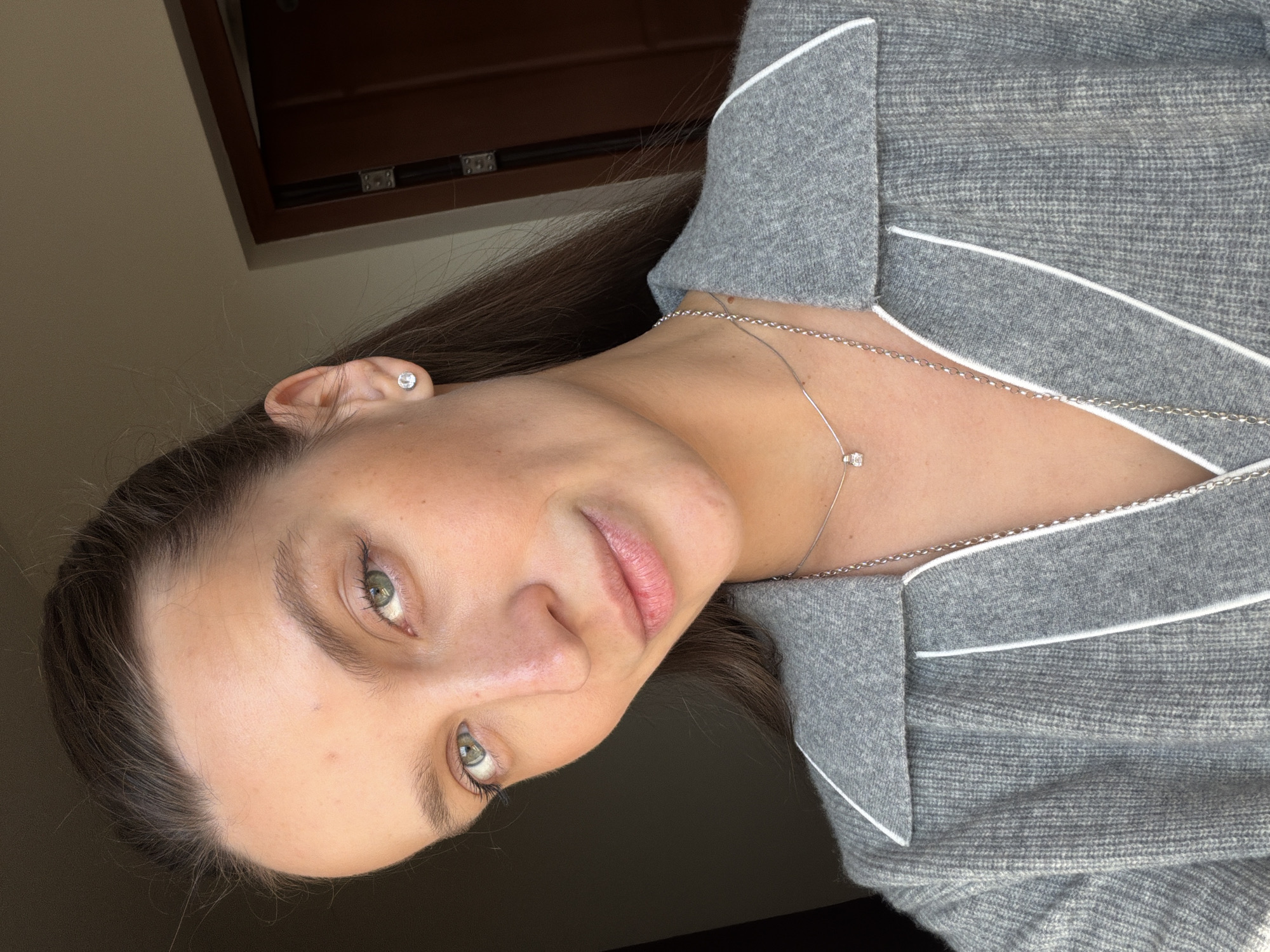 A close-up portrait, oriented diagonally, features a young woman with fair skin and striking green eyes. Her dark brown hair is pulled back, and she looks directly at the camera with a calm, neutral expression and a slight, soft smile. Visible freckles dot her cheeks. She wears a small, clear stud earring in her right ear and a delicate silver chain necklace with a small, clear gemstone pendant. Another thin silver chain is partially visible.
She is dressed in a soft grey top, possibly loungewear or pajamas, characterized by its textured fabric and prominent white piping along the collar and lapel edges. The background is softly blurred, revealing a warm-toned indoor setting. A dark wooden structure, possibly a window frame or furniture, is visible in the upper part of the image. The lighting is soft and natural, suggesting daytime. There is no visible text.