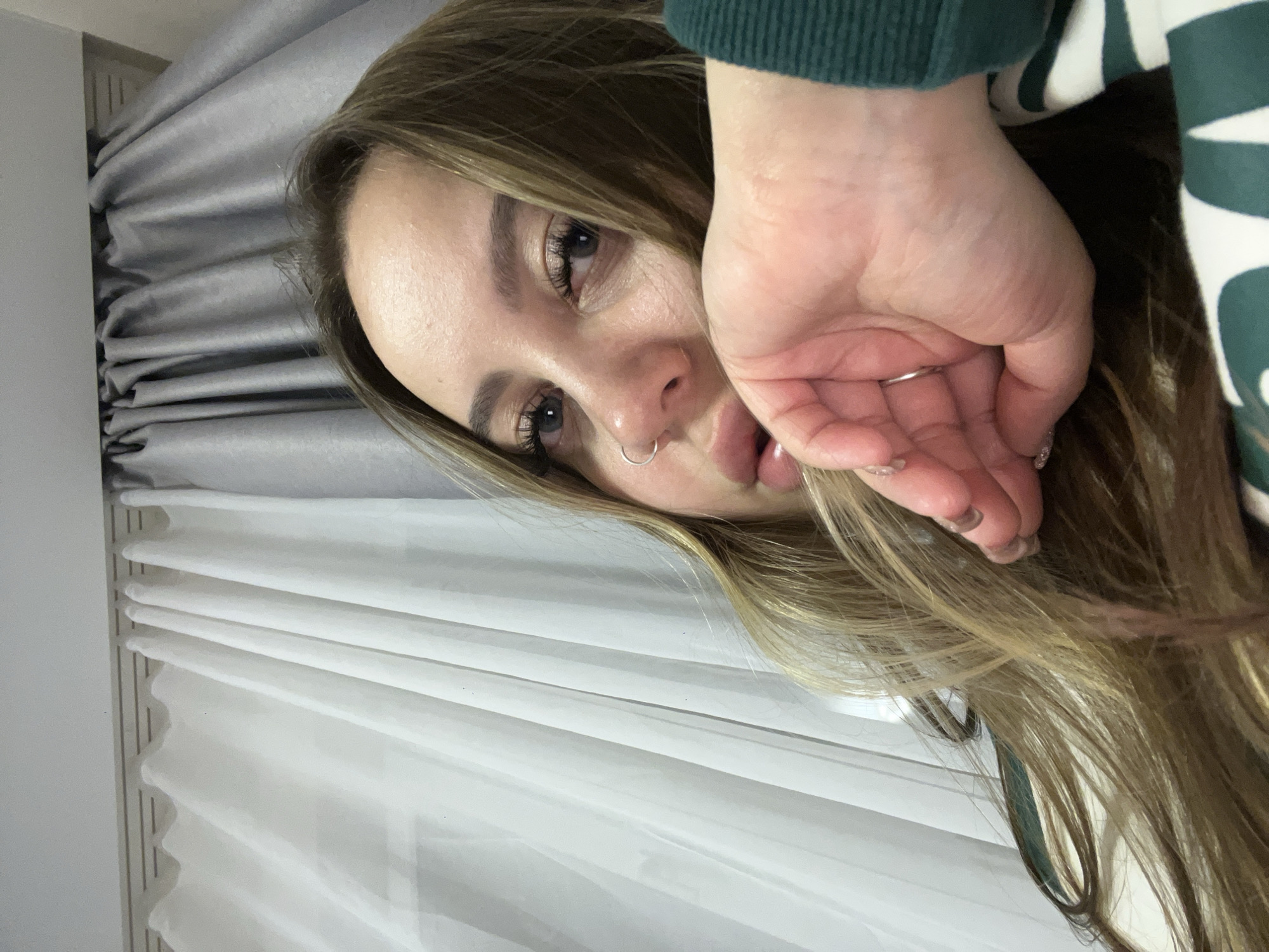 A young woman with long, light brown hair poses for a close-up selfie. She has fair skin, visible eyelashes, and a silver nose ring. Her right hand is raised, partially obscuring her face, with a silver ring visible on her ring finger. She wears a green and white patterned top. The background features layered, white and gray curtains or blinds. This picture's location in Krasnodar, Russia is not determinable from the picture.