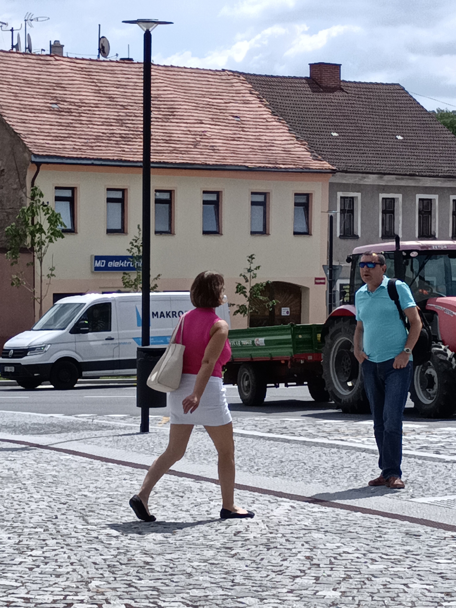 The image shows a street scene in Dalovy, Czechia, on a bright, partly cloudy day. In the foreground, a woman with brown hair, wearing a pink sleeveless top and a white skirt, is walking away from the camera. She is carrying a light-colored tote bag over her right shoulder. To her right, a man wearing a light blue polo shirt, blue jeans, and sunglasses is standing and looking towards the left. He has a backpack slung over his left shoulder. In the background, a red tractor is parked next to a green trailer. To the left of the tractor, a white van with the logo "MAKRO" is visible. Behind the van and tractor, there are buildings with tiled roofs. A street lamp is positioned in the middle ground, to the left of the people. The ground is paved with cobblestones.