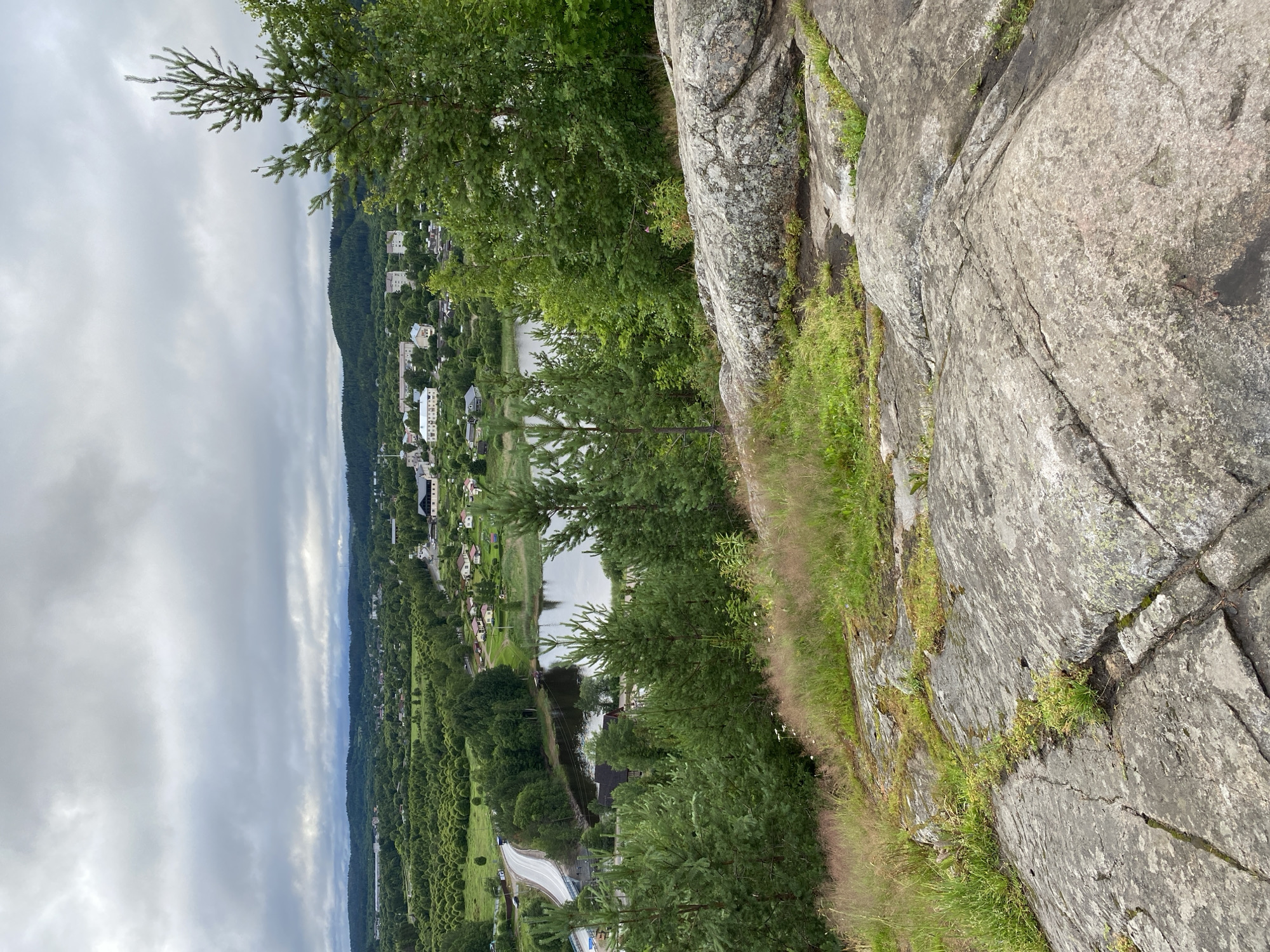 The image presents a scenic overlook of Lakhdenpokhya, Russia, on a cloudy day. In the foreground, large, weathered grey rocks covered in patches of green moss and grass dominate the scene, with a scattering of dry, tall grass. Behind the rocks, a lush green landscape unfolds, characterized by rolling hills dotted with numerous evergreen trees and deciduous bushes.
In the mid-ground, a calm, dark body of water, likely a lake or river, reflects the overcast sky. Along its edge, a small cluster of buildings, appearing to be houses and perhaps some larger structures, can be seen nestled amongst the trees. A paved road winds its way from the left side of the frame, crossing over a bridge and leading towards the settlement. Further into the distance, more buildings of varying sizes, including apartment blocks, are visible, forming the heart of the town. The horizon is defined by a line of dense, dark green forested hills under a thick, grey, cloud-filled sky. No people are visible in the image.