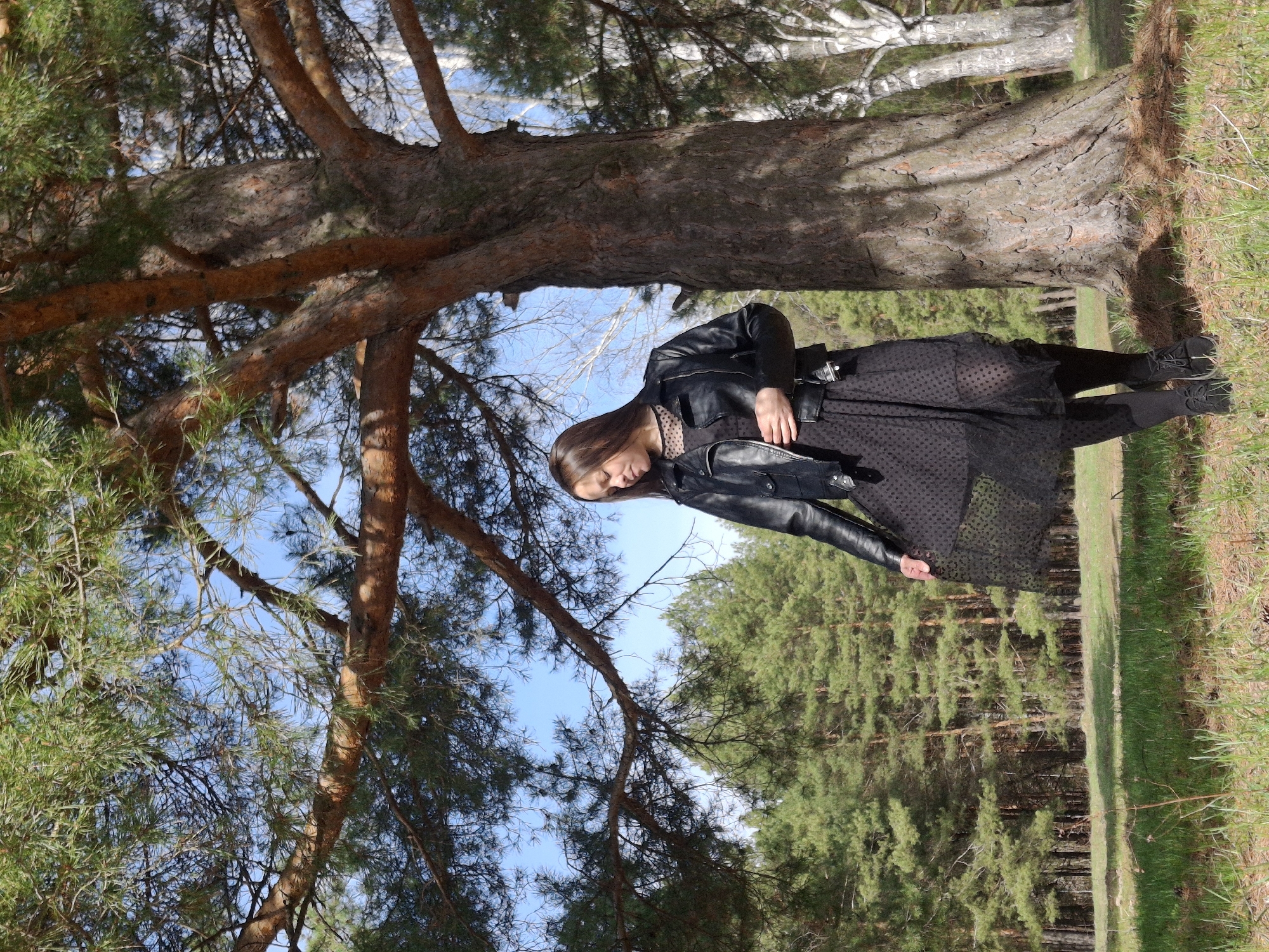 A full-length photograph depicts a woman standing beside a tree in what appears to be a wooded area. The location is the city of Chaikovsky, Russia. The woman is wearing a black leather jacket over a patterned dress, black tights, and black shoes. She is looking down. The large tree has a thick trunk, and the background includes numerous other trees. The ground is covered with grass and some dry vegetation. The sky is visible through the branches.
