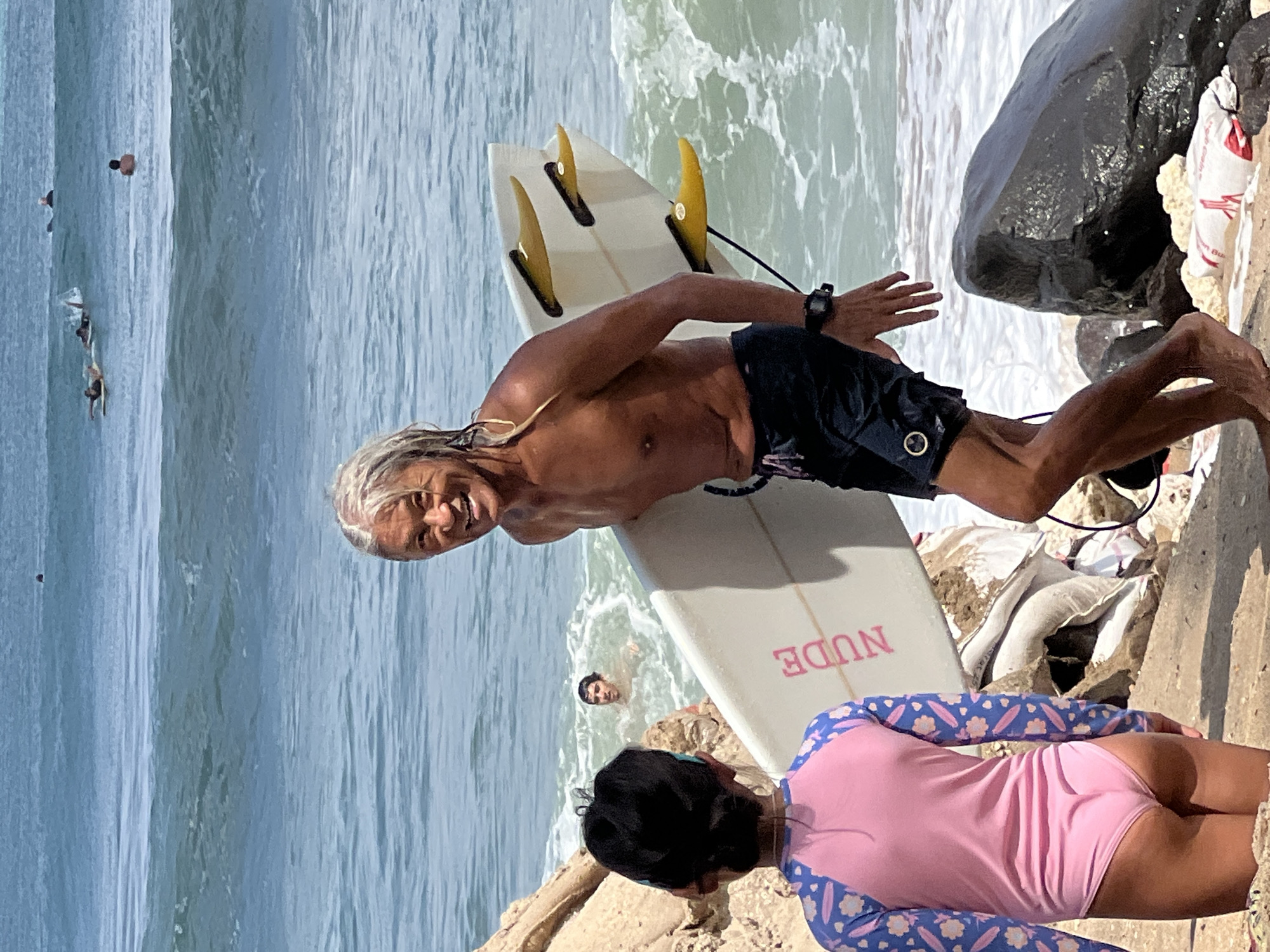 A surfer with long, gray hair and a muscular build is walking on a sandy beach in Kuta, Indonesia, carrying a white surfboard with yellow fins. He is smiling at the camera. A young girl in a pink and blue swimsuit with a floral pattern is standing in front of him, facing away from the camera and looking towards the ocean. In the background, several other surfers are in the water, some on their boards and others swimming. The ocean is blue with white waves. The beach is lined with rocks and some debris. The scene appears to be during the day, with bright sunlight.