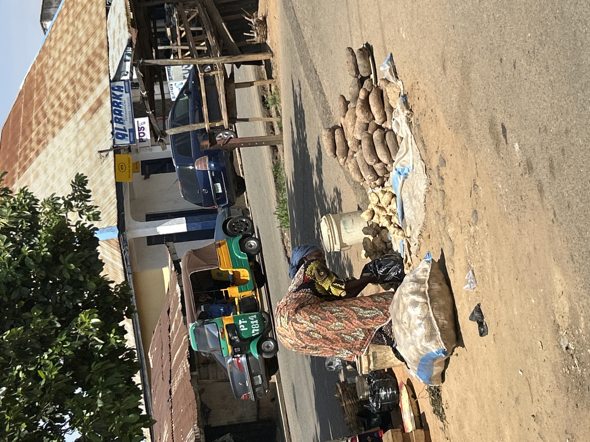 A street scene in Jos, Nigeria, shows a woman seated on the roadside near a pile of yams and smaller white objects, possibly potatoes. She is wearing a colorful patterned dress and a dark head covering. Behind her are bags and other merchandise. To the left, a green and yellow "keke napep" (motorized tricycle taxi) is parked next to a silver car. Further back, a blue car is parked in front of a building with a sign "ALBARKA." The building has a corrugated metal roof. A tree occupies the left side of the frame. The road appears to be paved and is mostly devoid of traffic.