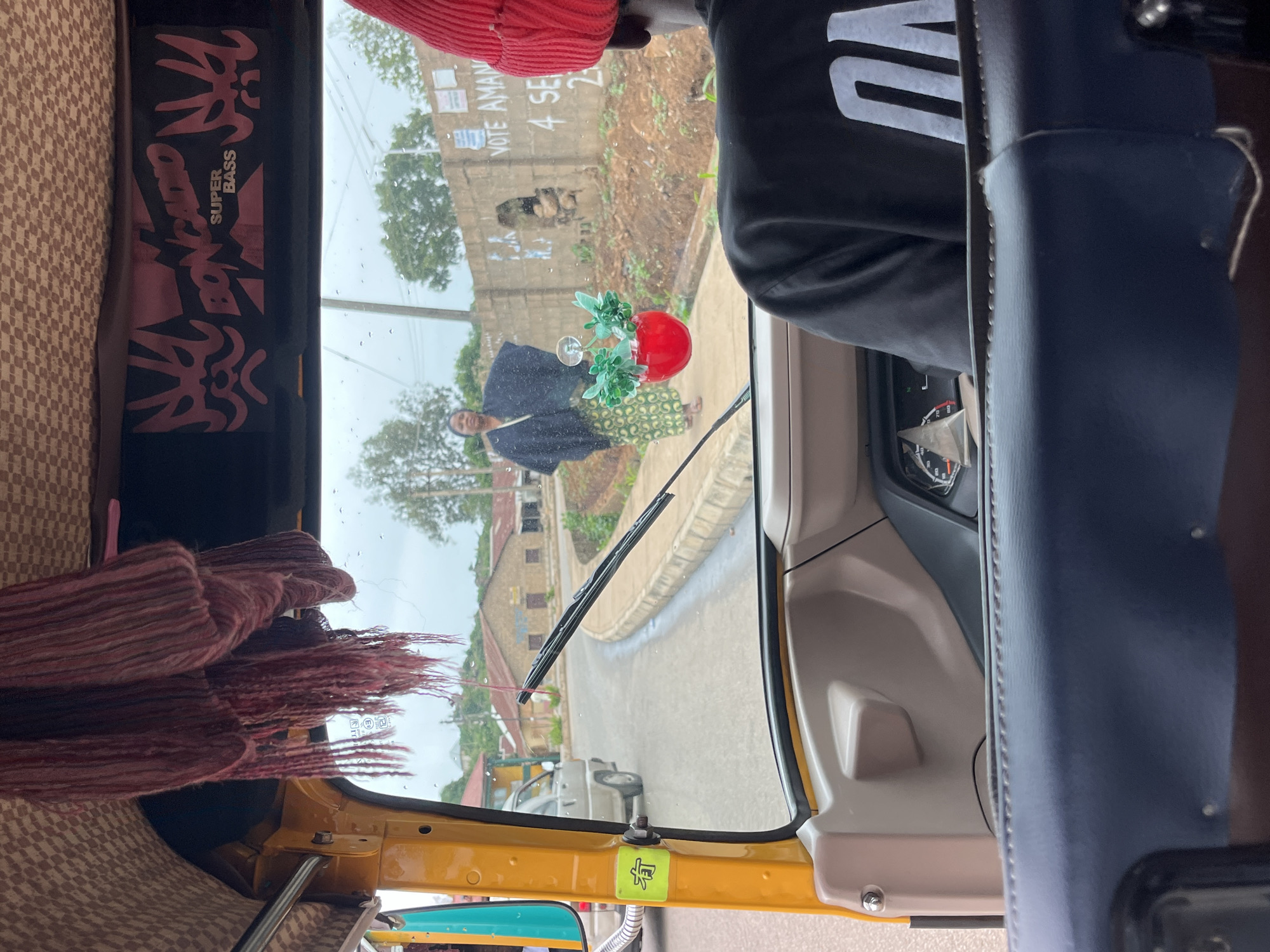 This is a photo taken from inside a yellow auto-rickshaw, looking out through the windshield. The scene appears to be a street in Bukuru, Nigeria, possibly during the day. The weather seems overcast or rainy, as evidenced by the wet windshield with visible raindrops.
In the middle ground, outside the auto-rickshaw, a woman is walking, carrying a red pot with some green decorative elements. She is wearing a dark-colored garment and a patterned skirt. Behind her, a building with a weathered facade and signs is visible, suggesting an urban or semi-urban setting. One of the signs appears to be a campaign poster with the text "VOTE AMAN" and "4 SE...". The ground is wet, and a street curves away into the distance.
Inside the auto-rickshaw, the interior is partially visible. To the left, there is fabric with a patterned texture and some red fringed material. A black banner with pink lettering is attached to the interior, featuring the text "SUPER BASS" and other stylized graphics. To the right, a dark blue seat or panel is prominent. The driver's side of the auto-rickshaw is also visible, with a steering wheel and dashboard instruments partially in view. A person's shoulder and arm, wearing a black t-shirt with white lettering that appears to say "VNO", are seen on the right side of the frame, suggesting the photographer is a passenger or the driver. A yellow sticker with a symbol resembling a person is visible on the yellow frame of the auto-rickshaw. The overall impression is a glimpse of daily life on a street in Nigeria, viewed from the perspective of someone traveling in an auto-rickshaw.