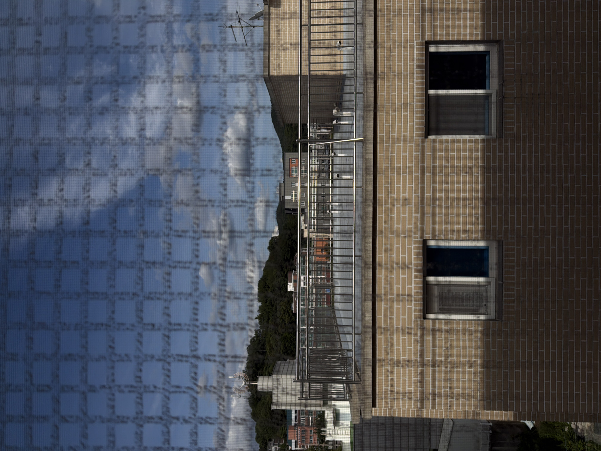 The image shows a view of the city of Dongducheon-si, South Korea, from an elevated position, looking out over rooftops. In the foreground, there is a brick building with two windows. The building appears to be residential or commercial. Above this building, there is a rooftop with a metal railing and some piping. Beyond the rooftop, a cityscape unfolds with various buildings of different colors and heights, including some with red accents. Further in the background, a lush green hillside is visible, topped with trees. The sky is blue with scattered white clouds, and the entire scene is overlaid with a fine grid pattern, suggesting a screen or a textured surface in front of the camera. The lighting suggests it is daytime, possibly late morning or early afternoon, with some shadows cast on the brick building. There are no people visible in the image.