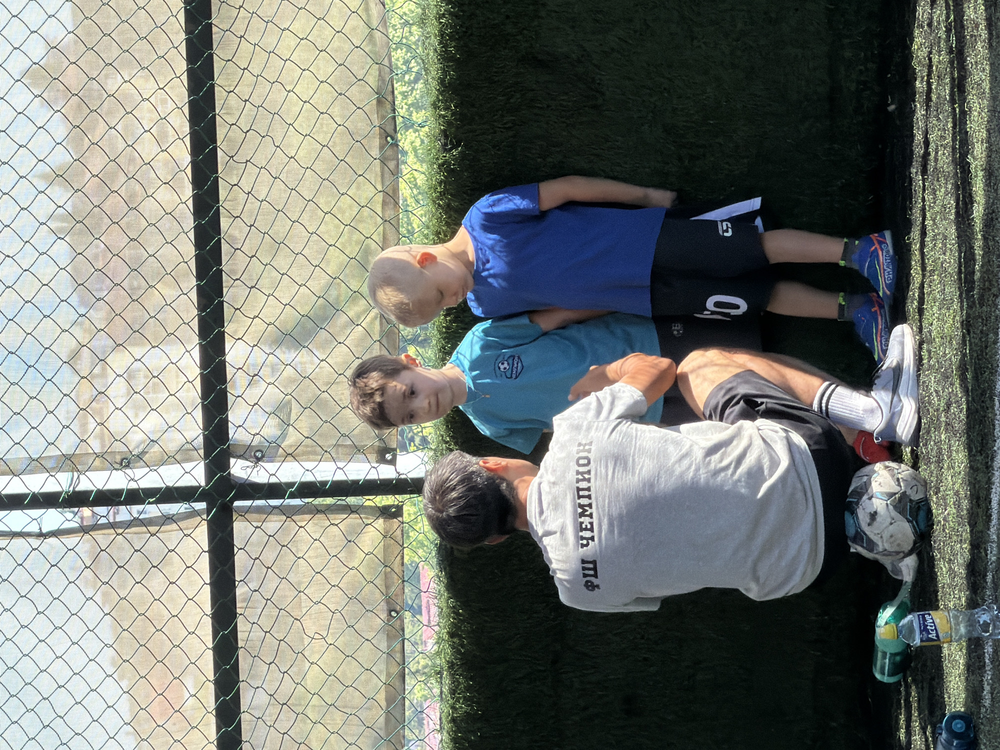 The image depicts an outdoor scene at a soccer training facility in Sochi, Russia. An adult male, viewed from the back, is crouched on an artificial turf field, engaging with two young boys standing in front of him.
The adult male wears a light grey short-sleeved t-shirt with "ФШ ЧЕМПИОН" printed in dark grey Cyrillic letters across the upper back, which translates to "Football School Champion." He has dark hair, black athletic shorts, white socks, and grey Nike athletic shoes. Beneath him, on the artificial turf, is a deflated black and white soccer ball. To his left, a green water bottle and a clear "Active" brand water bottle with a yellow cap lie on the turf, alongside other dark objects.
To the left of the adult is a young boy with light brown hair, wearing a light blue short-sleeved t-shirt with a darker blue logo featuring a soccer ball and text (likely "ЧЕМПИОН") in Cyrillic, and dark shorts. To the right of the adult is another young boy with short blonde hair, wearing a royal blue short-sleeved t-shirt and black athletic shorts with a white '0' and "CS" logo on the left leg. He wears blue athletic shoes with orange and pink accents, and the word "PHANTOM" is visible on the side of one shoe. Both boys are looking towards the adult.
The background features a dark green chain-link fence, supported by a dark horizontal bar and a white vertical post. Behind the fence, a blurred view of trees, foliage, and some distant structures under a bright sky is visible, indicating a sunny day. The lower portion of the background is a green artificial turf wall, matching the ground. The textual evidence on the adult's shirt, "ФШ ЧЕМПИОН," identifies the location as likely a training ground for the "Football School Champion" in Sochi, Russia.