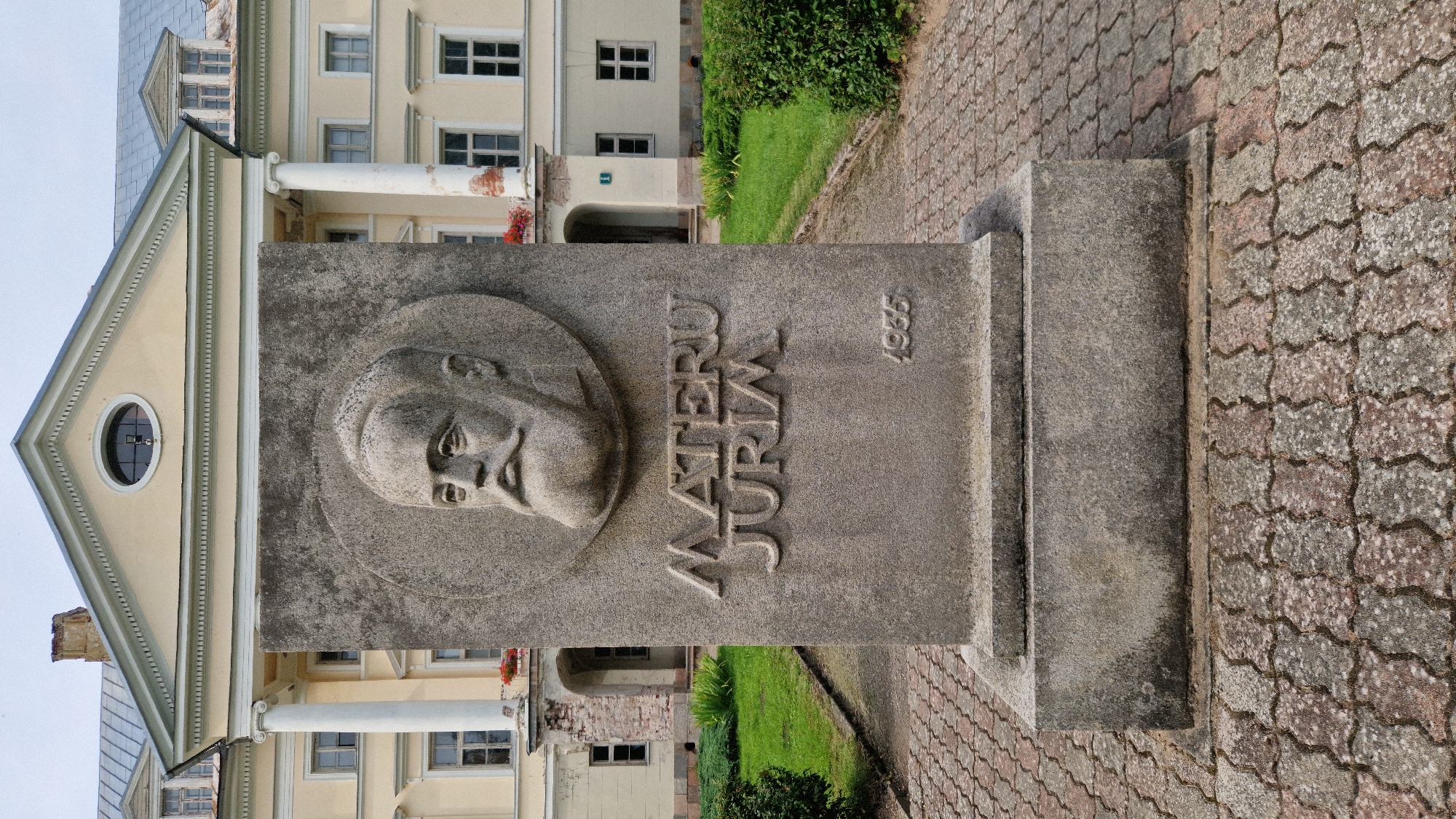 In Kazdanga, Latvia, a stone monument stands in front of a pale yellow building with white columns. The monument features a bas-relief portrait of a man with a beard and mustache, enclosed in a circular border. Below the portrait, the inscription "MATERU JURIM" is carved, followed by the year "1935" to the right. The monument is placed on a paved walkway made of interlocking bricks. Green grass and shrubs are visible on either side of the walkway. The building in the background appears to be a historical structure, possibly a manor or public building, with multiple windows and a light-colored roof.