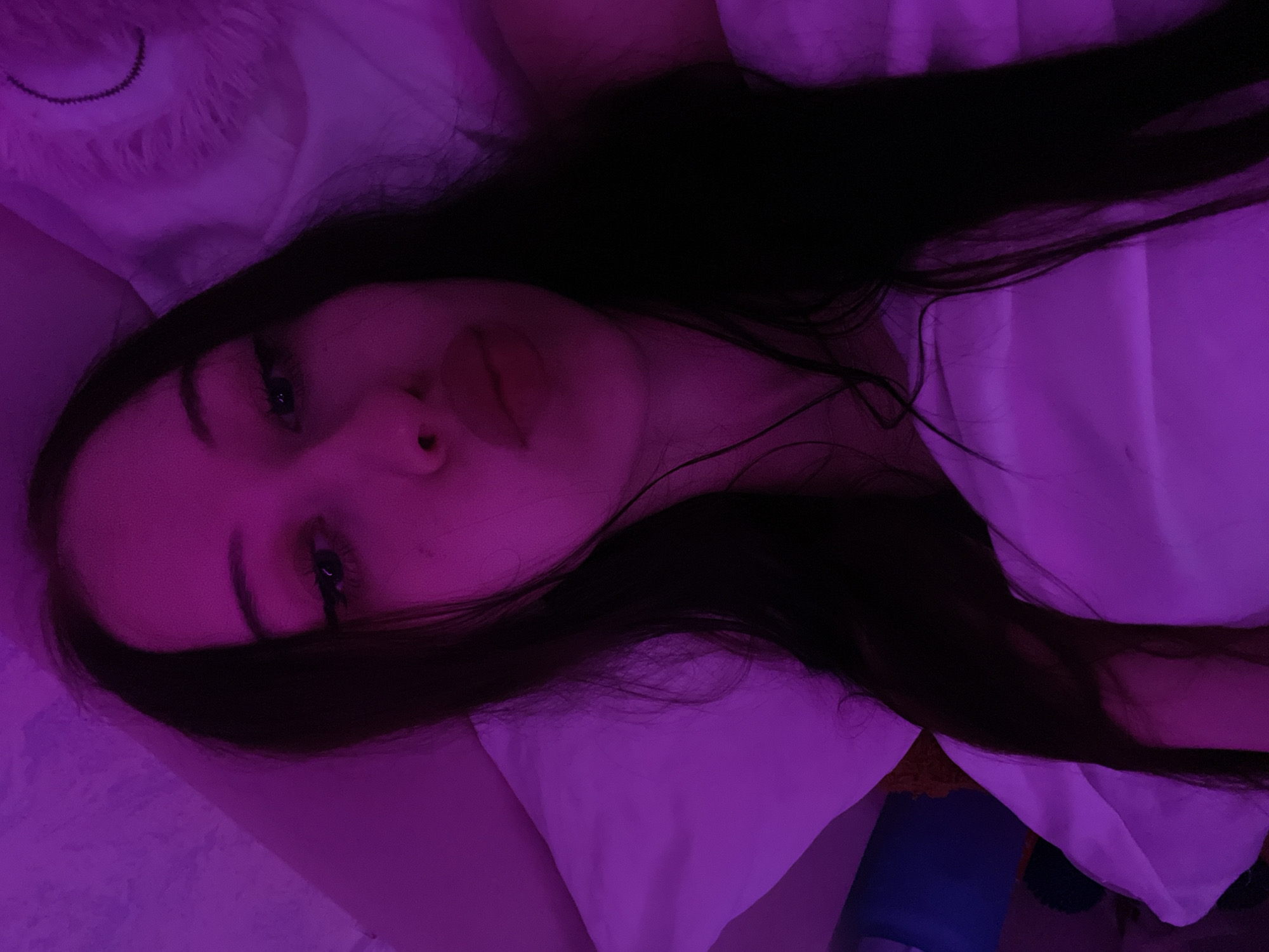 The image shows a close-up, overhead view of a young woman with long, dark hair lying on a white pillow. She is looking up and to the left, with her lips pursed. The room is lit with purple light. A fluffy, white object, possibly a stuffed animal or a pillow, is visible to the right of the woman's head. There is a hint of a blue object in the background on the left. There is no information in the image to suggest the location is Votkinsk, Russia.