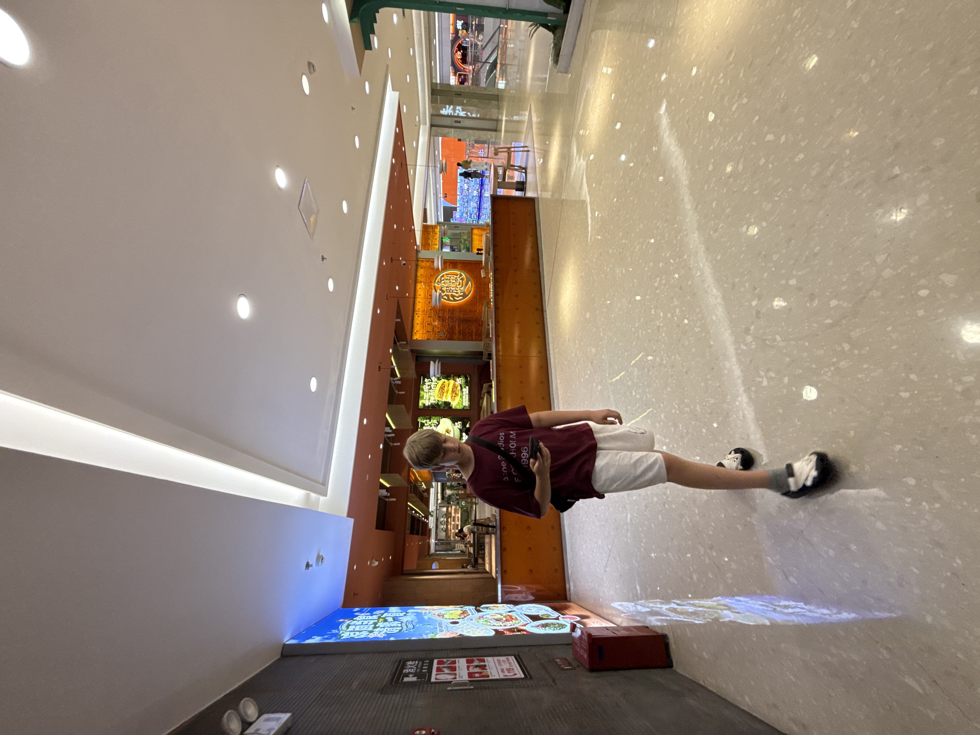 A young Caucasian male with blond hair is standing indoors, facing slightly to his right while looking down at a smartphone in his hands. He is wearing a burgundy t-shirt with white lettering and white shorts, paired with dark sandals and gray socks. A black strap from a bag is visible over his left shoulder.
The setting appears to be a modern indoor commercial space, possibly a shopping mall or a food court in Huizhou, China. The floor is a polished light-colored terrazzo with scattered reflective elements. To the left of the individual, a large, illuminated digital display board is mounted on a dark grey textured wall, featuring colorful images and Chinese text. A red fire alarm box is situated at the base of this wall.
In the background, a brightly lit food establishment is visible, with an orange counter and glass displays showcasing food items. Various tables and chairs are set up in front of the establishment. Further in the background, there are escalators and other retail spaces, indicative of a large commercial complex. The ceiling is a modern design with recessed lighting and illuminated paneling. The overall ambiance is well-lit and spacious.