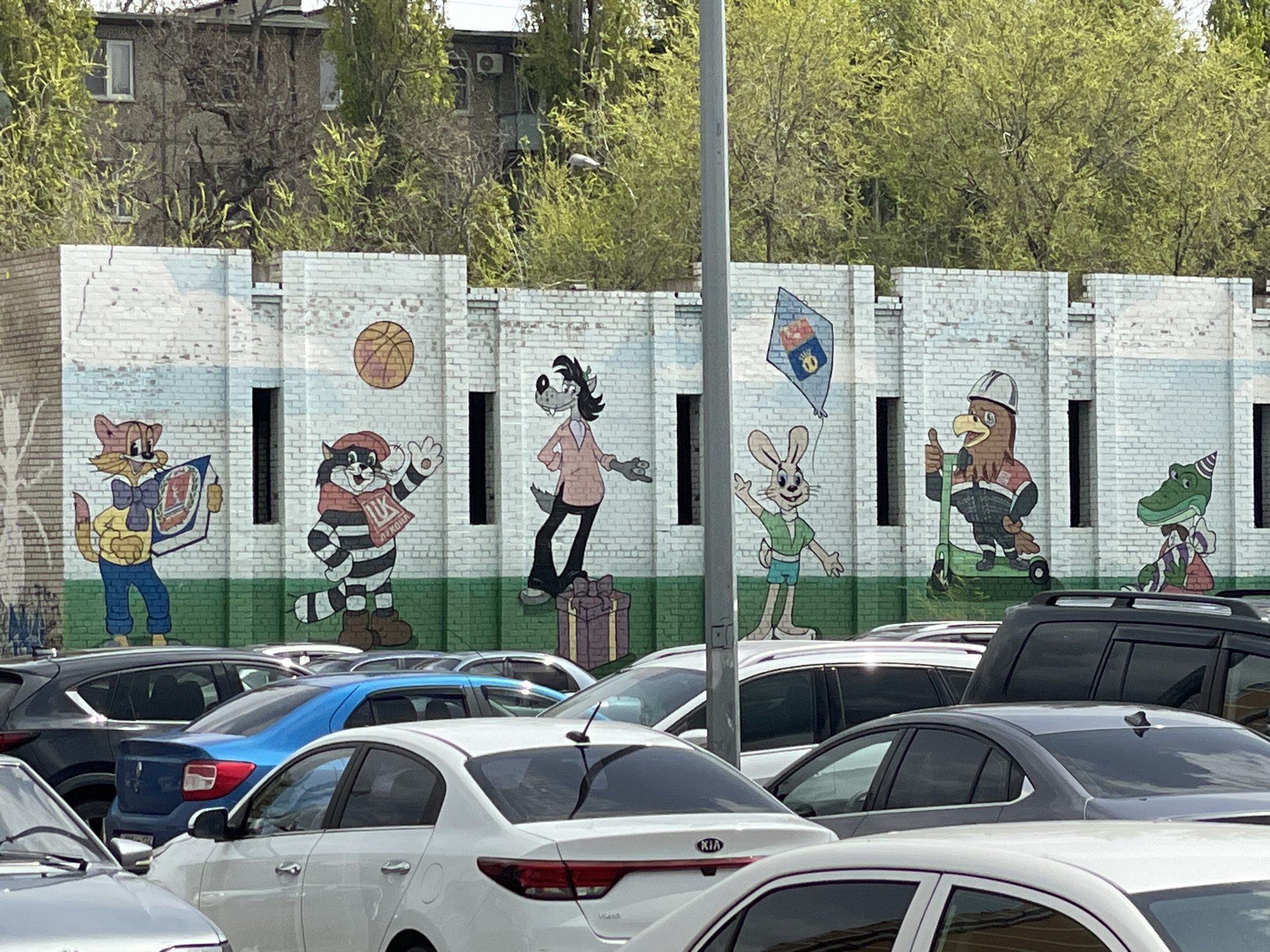 An outdoor, brightly lit scene features a colorful mural painted on a low, white-painted brick wall. The mural depicts six anthropomorphic cartoon characters. From left to right: a cat-like character in a yellow shirt holds an open book; a striped feline character, resembling a raccoon or cat, in a red hat waves, with "ЛУКОЙЛ" (LUKOIL) visible on its sleeve; a wolf character in a pink jacket stands on a wrapped gift box; a rabbit character in a green shirt holds a kite featuring a crown emblem; an eagle character in a hard hat gives a thumbs-up with a tool; and a crocodile character in a striped shirt sits on a scooter. A basketball is painted in the air above the second character. The characters appear cheerful and energetic.
In the foreground, a busy parking lot is filled with numerous parked cars, including sedans and SUVs of various colors, mostly seen from their rear ends. A tall grey utility pole partially obscures the central part of the mural.
Behind the mural, trees with vibrant, light green foliage suggest the season is spring or early summer. Through the gaps in the trees, parts of a multi-story, utilitarian building are visible in the background, hinting at an urban or residential environment. The clear daylight indicates sunny weather. The art style of the characters, particularly the wolf and rabbit, is reminiscent of Soviet-era animation (e.g., "Nu, pogodi!"), and the visible "LUKOIL" text strongly suggests the location is in Russia or another post-Soviet country.