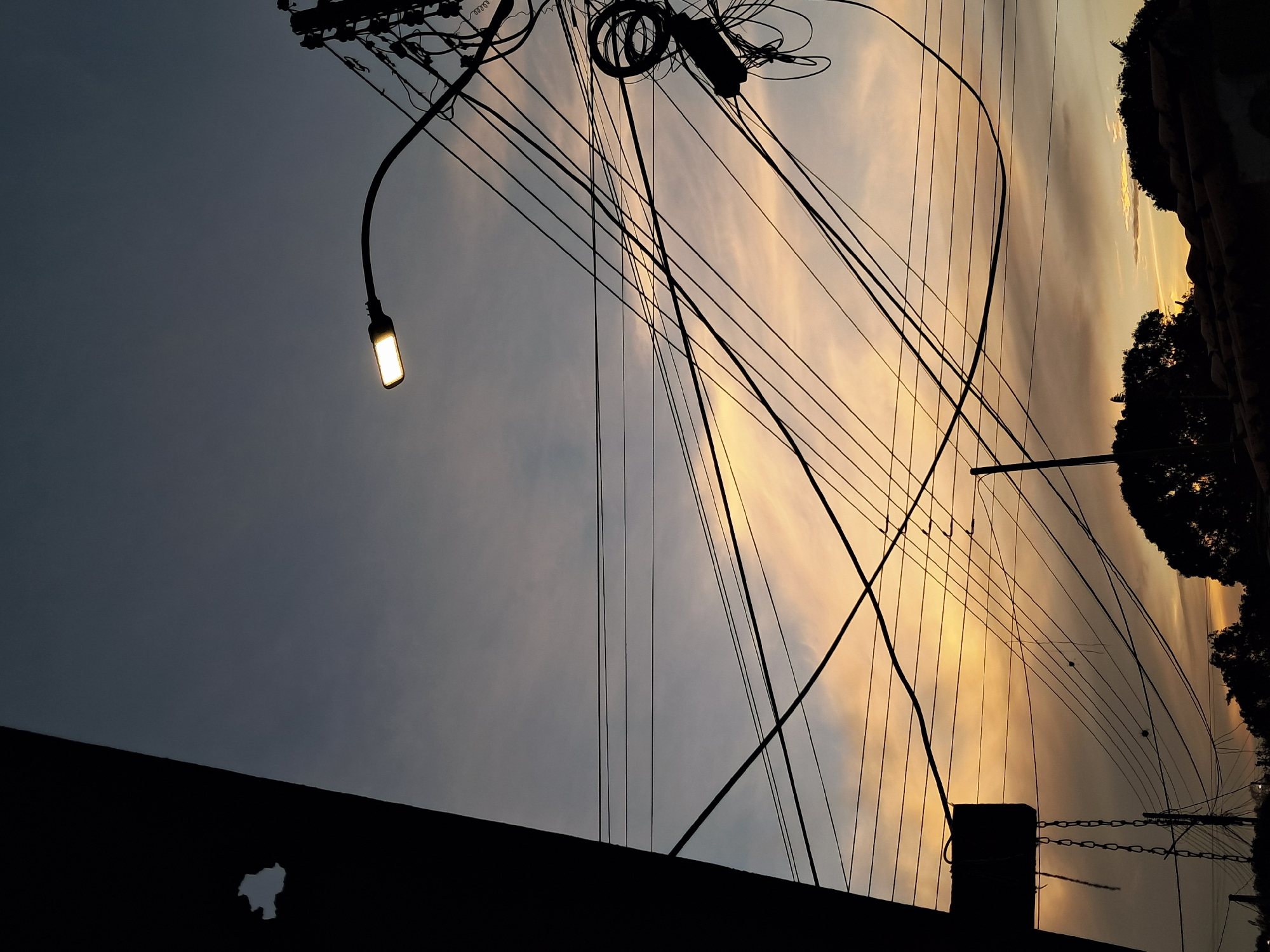The image depicts a twilight scene in Juan Griego, Venezuela, dominated by utility infrastructure. An illuminated street lamp on a curved pole is visible in the upper left corner, while numerous electrical wires crisscross the sky. Silhouetted trees and buildings are present along the right edge, and more silhouetted structures are at the bottom. The sky exhibits a gradient of color, ranging from a darker blue-gray on the left to a lighter orange-yellow on the right, indicating either sunrise or sunset.