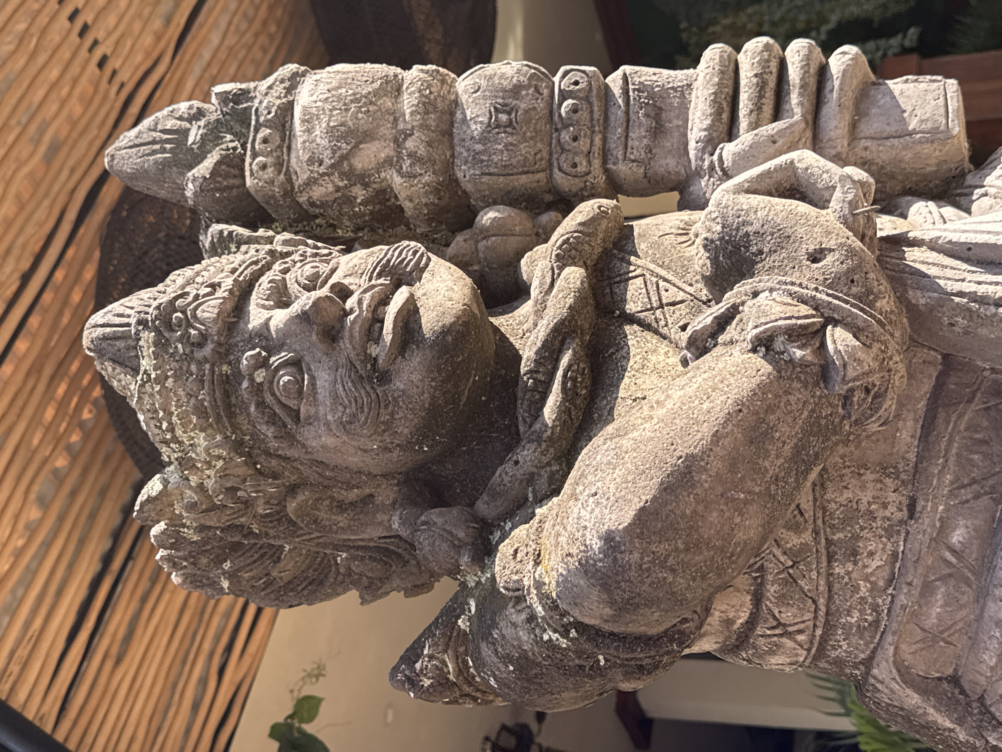 A stone statue of a deity, possibly a guardian, stands outside under a thatched roof structure. The statue is intricately carved with detailed facial features, including bulging eyes and a mustache. It wears a crown and jewelry. The deity holds an object in its left hand and rests its right arm on a cylindrical pillar. The stone is weathered and shows signs of age, with some moss and lichen growth. The background suggests an outdoor setting in a tropical or subtropical location, consistent with Ubud District, Indonesia. The lighting indicates it is daytime, with sunlight casting shadows.