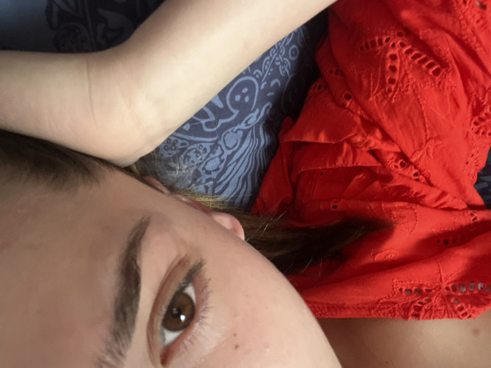 The image provides a close-up, upward-tilted perspective of a single human subject. The subject's head is angled, prominently displaying the right eye, which is brown, and a dark, shaped eyebrow. Fair skin is visible on the forehead, temple, and a portion of the cheek. Long, dark hair is present near the subject's shoulder. The subject wears a bright red garment, possibly a top or dress, featuring intricate eyelet embroidery and cutouts. The subject's right arm is flexed, positioned near the head. The individual is situated on a blue textile surface adorned with an intricate pattern of lighter blue and white swirling motifs, consistent with bedding or upholstery. The exact location within Ancenis, France, is not identifiable from the visual information provided.
