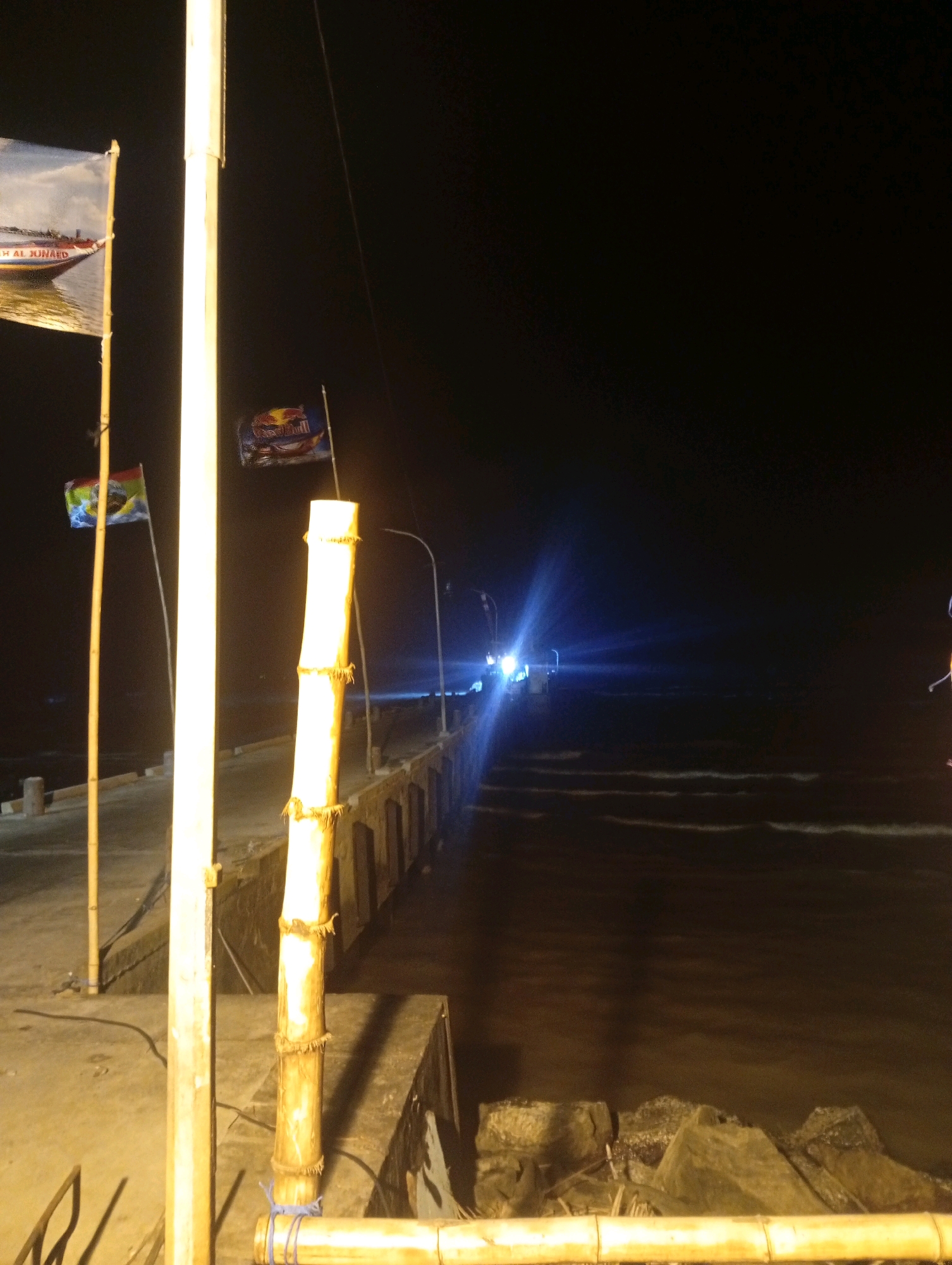 The image depicts a night scene at the Banowan, Indonesia harbor. A concrete pier extends into the dark water, illuminated by artificial lights. Several bamboo poles are visible, some tied with blue rope, and a tall, bright light pole stands prominently in the foreground. Two flags are mounted on poles to the left of the light pole; one is a large rectangular flag with an image of a boat and text that appears to say "AL JUNAID," and the other is a smaller, triangular flag with colorful designs. Further down the pier, another small flag, a Red Bull logo, is visible. In the distance, lights from a boat or structure create a strong blue-white glare on the water's surface. The water exhibits light ripples, suggesting calm conditions. Rocks are present along the edge of the pier in the foreground.