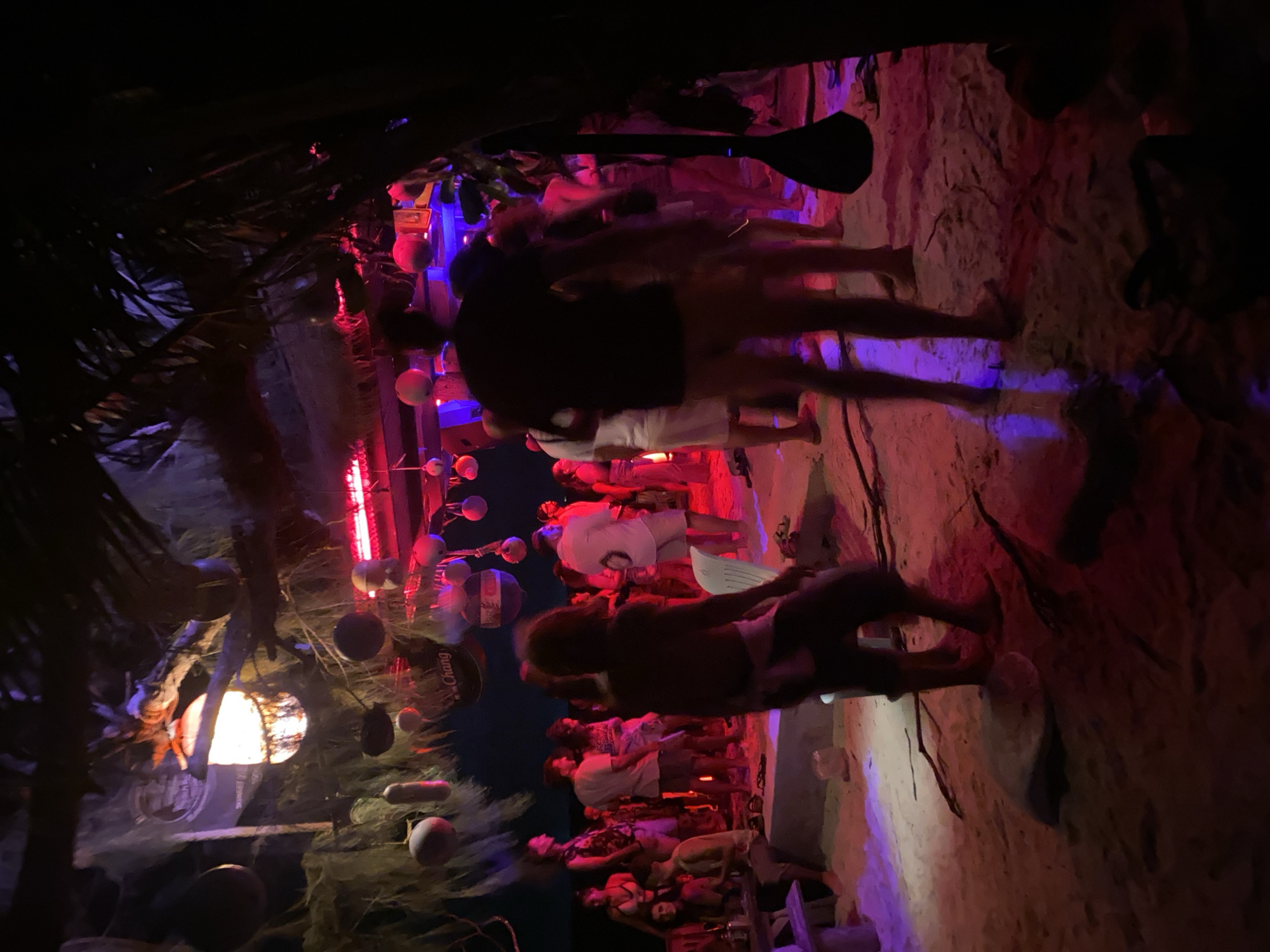 The image shows a bustling beach bar scene at night in Baan Sri Thanu, Thailand. Several people are present, some walking around, others standing in groups. The environment includes a sandy beach, illuminated by red and purple lights. Overhead, there's a thatched roof structure adorned with various decorative items, including round balls, Chang beer signs, and what appears to be a paddle. The overall atmosphere suggests a lively and social gathering.