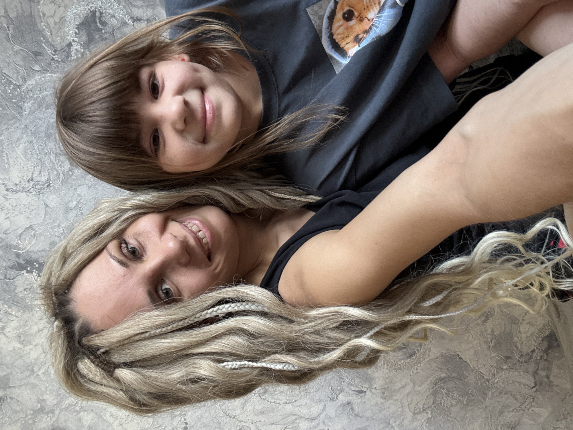 A photograph depicts two individuals lying on what appears to be a textured gray surface, possibly a floor or bedding. The individual at the top of the frame is a child with brown hair and bangs, wearing a gray shirt with an image of a cat on it. They are smiling towards the camera. Below them is an adult with long, blonde hair styled with braids. The adult is wearing a black shirt and also smiling at the camera. The setting is ambiguous and does not clearly indicate the location in a city.