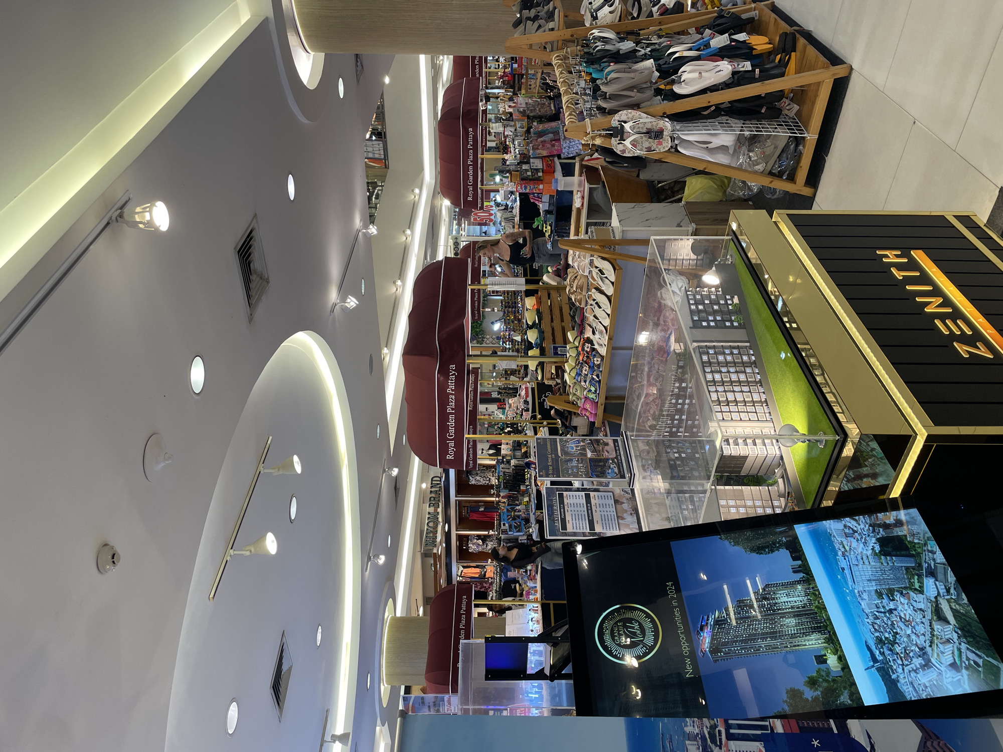 This image depicts the interior of a retail establishment in Royal Garden Plaza, Pattaya City, Thailand. The scene showcases a variety of goods, including footwear such as sandals and flip-flops displayed on wooden racks and shelves. Several retail stalls with red awnings are visible, with signage indicating "Royal Garden Plaza Pattaya" and "NIYOM BRAND". A woman is standing near one of the stalls, appearing to interact with merchandise. In the foreground, a display case features a miniature architectural model of a building complex situated on a green lawn, with accompanying promotional material for "Grand Solaire Noble" and the text "New opportunities in 2024." The environment is well-lit, with multiple ceiling lights and spotlights illuminating the space. The flooring appears to be tiled.