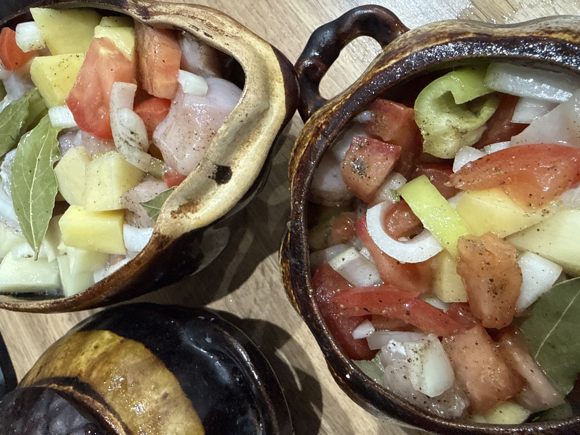 Two clay pots containing chopped ingredients are arranged on a wooden surface. The pot in the foreground is closer to the viewer and is filled with pieces of tomato, potato, onion, and what appears to be meat or fish, seasoned with pepper and bay leaves. The second pot, located behind and to the right of the first, also contains similar chopped ingredients. A dark, glazed ceramic pot is partially visible on the left side of the image. The lighting suggests an indoor setting, possibly a kitchen. There are no people visible in the image. The location in the city of Октябрьское, Russia is not discernible from the visual information provided.
