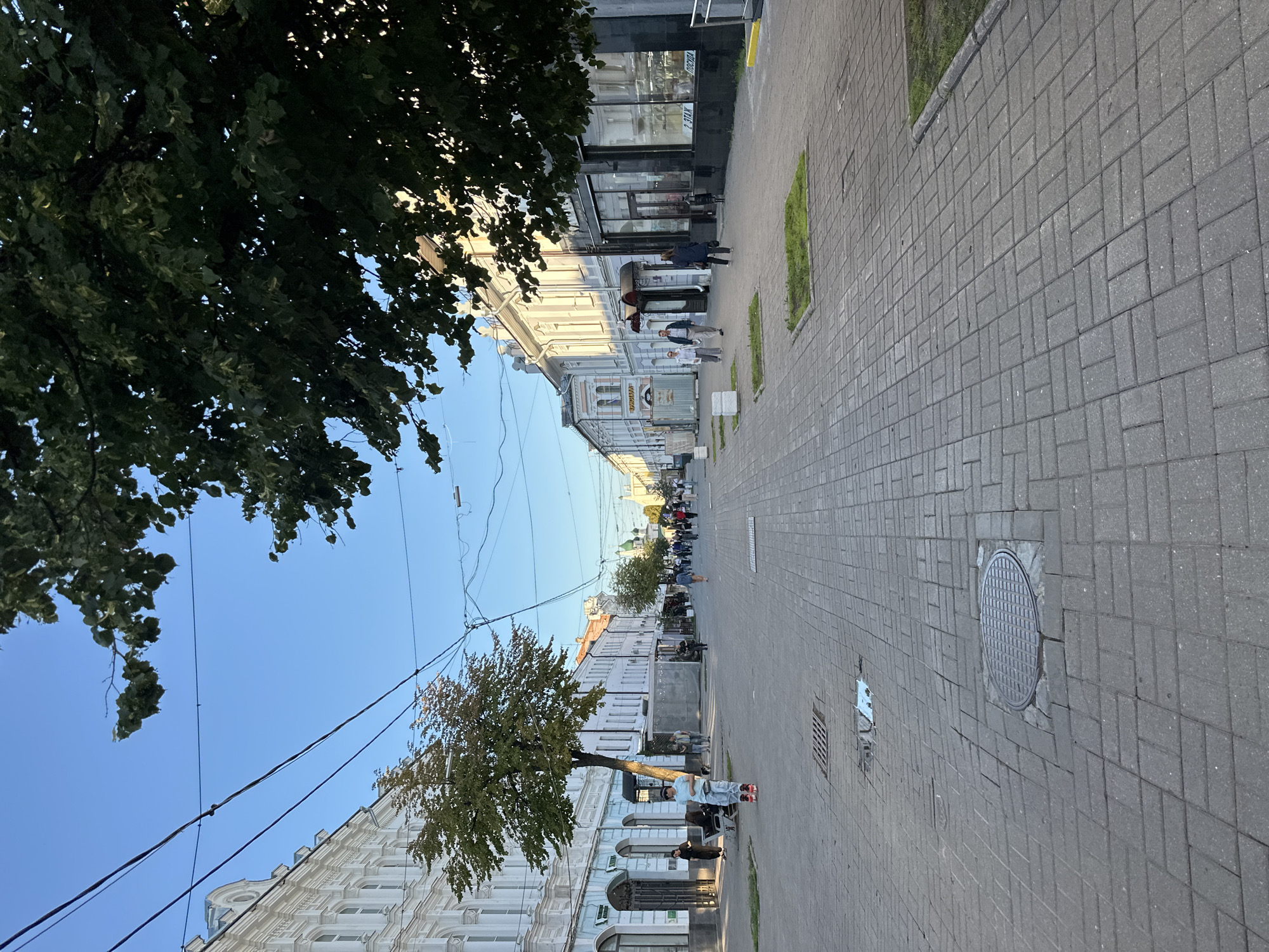The image shows a street scene in Feklino, Russia, with buildings lining both sides of a paved pedestrian walkway. A tree stands in the middle of the walkway, with its branches extending into the upper portion of the frame. Several people are walking along the street, some singly and some in groups. The buildings appear to be older, with traditional architecture. The sky is clear and blue, indicating a sunny day.