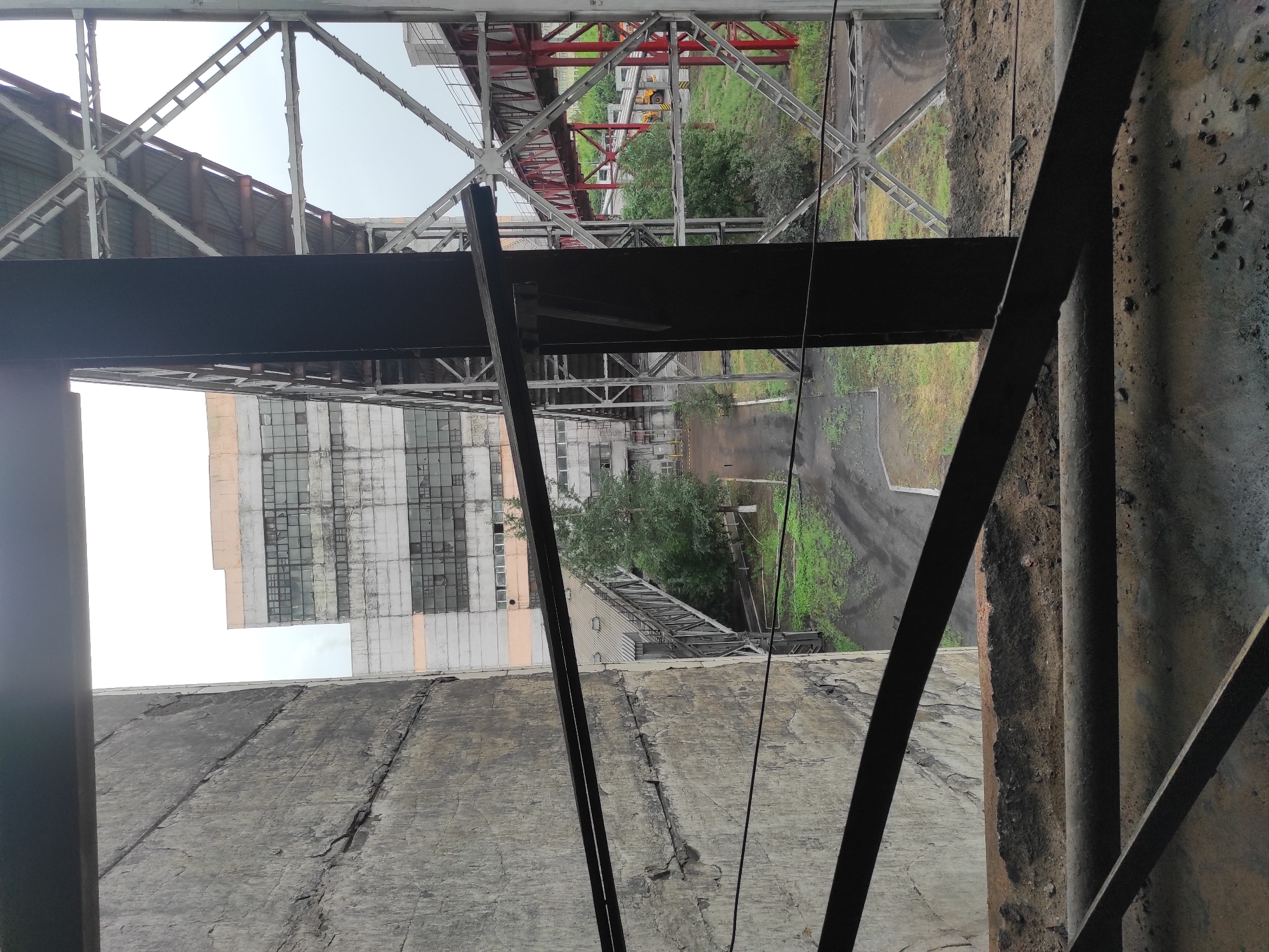 The image shows an industrial area, likely a former factory or processing plant, with a large concrete structure on the left and metal scaffolding and walkways in the foreground and background. A multi-story building with many windows, some broken, is visible in the distance. The ground is covered with dirt and debris, and there are some trees and vegetation. The sky is overcast. The scene suggests a derelict or abandoned industrial site. It is not possible to definitively identify this as being in Kyzyl, Kazakhstan, as there are no specific identifying markers or context provided within the image itself.
