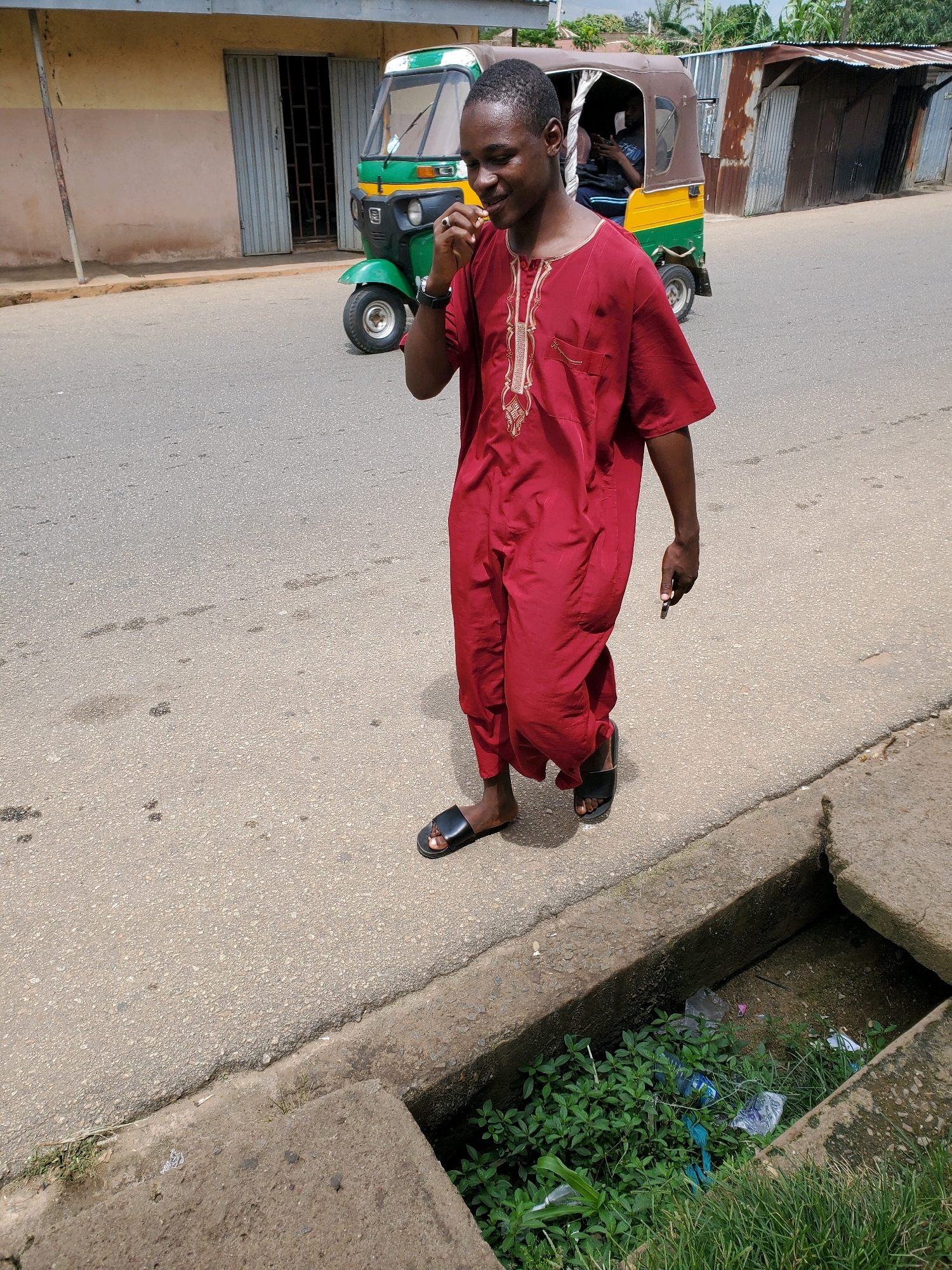 In Jos, Nigeria, a young man, dressed in a traditional maroon robe with intricate gold embroidery, walks along a paved road. He appears to be in motion, with his right hand touching his mouth, possibly eating or holding something. He is wearing black sandals. In the background, a brightly colored tuk-tuk, a three-wheeled auto-rickshaw, is parked. The tuk-tuk is predominantly green with a yellow front and brown trim on its canopy. A second person is visible inside the tuk-tuk. To the right of the tuk-tuk, a corrugated metal structure, likely a building or stall, stands with its doors closed. The road shows signs of wear, with scattered stains and a concrete drainage ditch running along the side. The ditch is partially filled with green vegetation and some discarded plastic bags, indicating a common urban environmental concern. The overall scene depicts a slice of everyday life on a street in Jos.