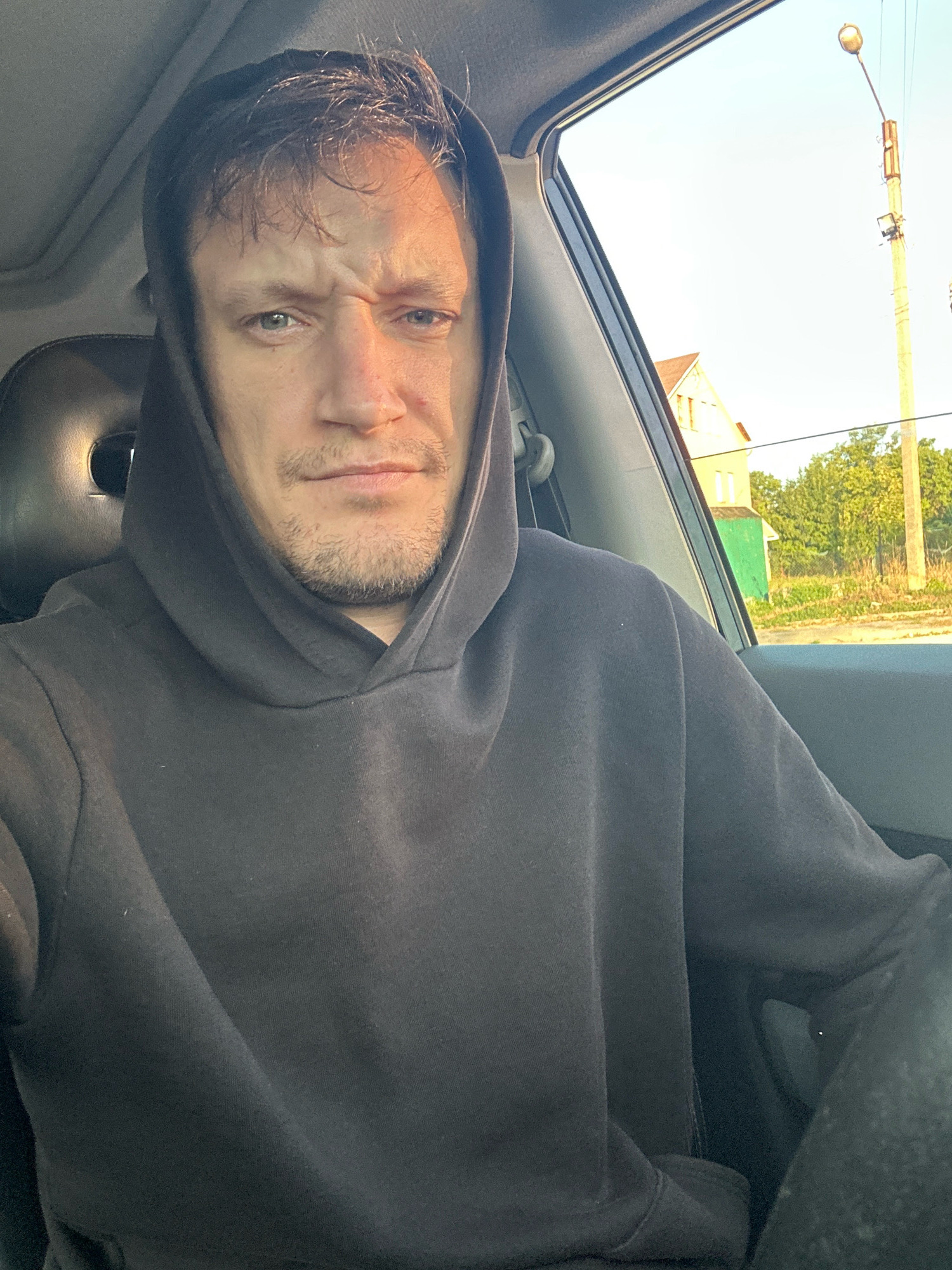 The image is a selfie taken from inside a vehicle, possibly a car, by a man wearing a dark hooded sweatshirt. He has light-colored eyes and short, dark hair. His expression appears serious, with furrowed brows and a slight frown. Sunlight is casting a shadow across the right side of his face.
The background visible through the car window shows a street scene with a yellow house, a green building, trees, and a tall street light. This scenery is consistent with a location in the city of Makiv, Ukraine, given the architectural styles and vegetation. The overall lighting suggests it is daytime, likely late afternoon or early morning, due to the angle of the sun. The interior of the car is dimly lit, with a black seat visible behind the man.