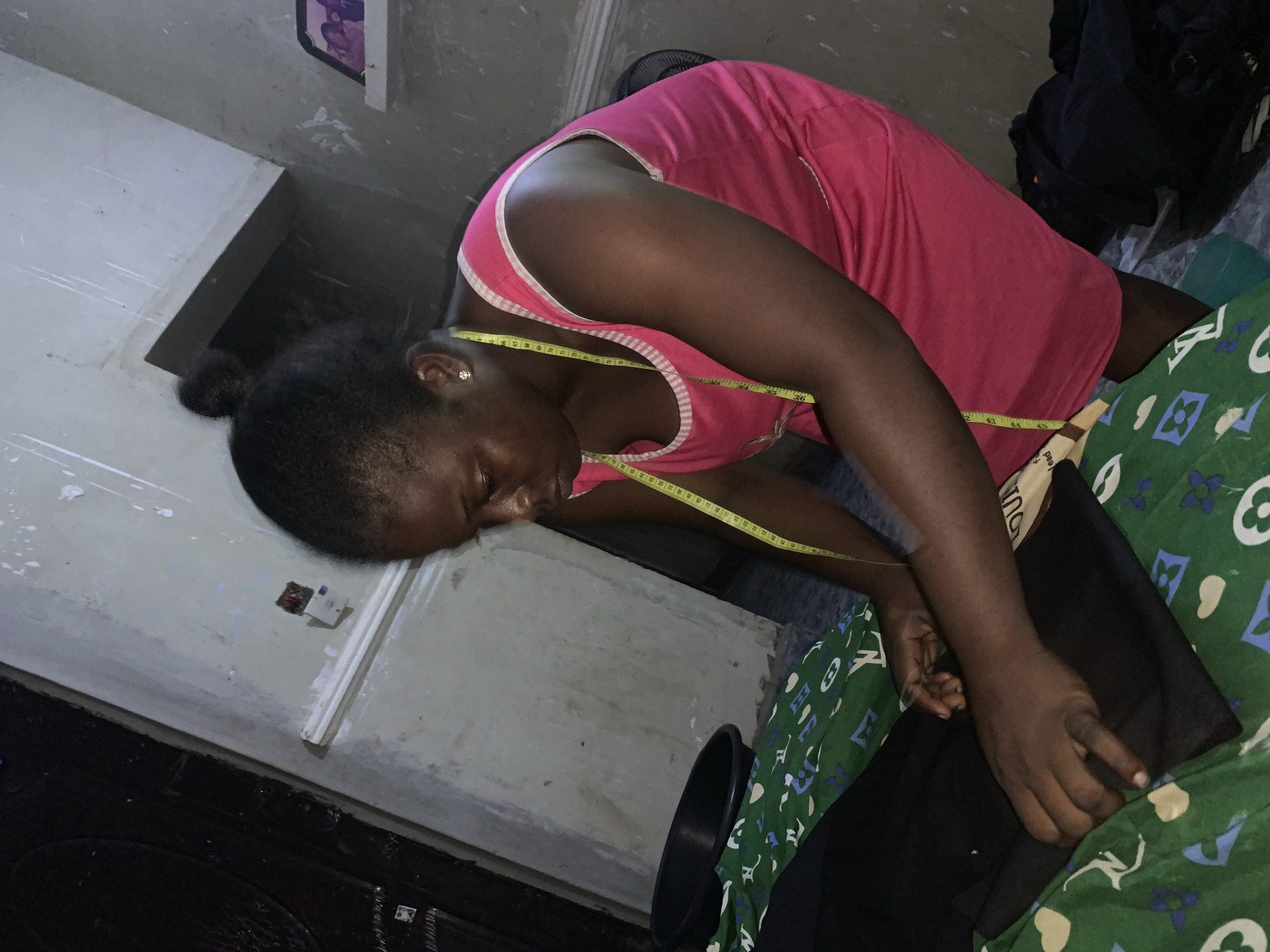 In a dimly lit room in Akwanga, Nigeria, a Black woman with her hair pulled into a bun is focused on a piece of black fabric spread across a green patterned surface. She wears a pink tank top with white trim and a yellow measuring tape draped around her neck. Her hands are engaged in handling the fabric, suggesting she is sewing or tailoring. A black basin is positioned to her left, and piles of dark clothing are visible in the background. The walls of the room appear worn and textured, with visible electrical conduits and a small shelf holding what appears to be framed photographs. The overall scene depicts a domestic or small workshop setting where sewing activities are taking place.