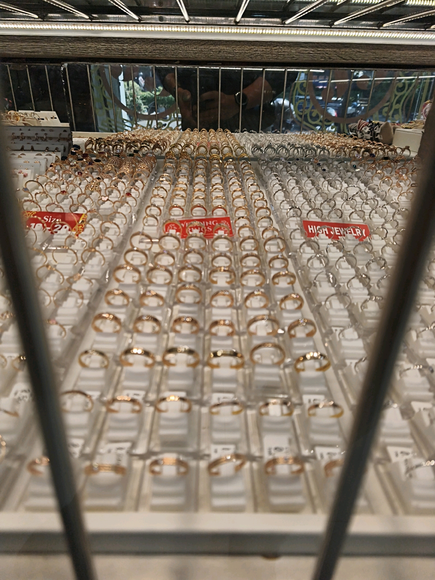 A close-up, slightly angled shot shows a display of rings in a jewelry store, likely in Surakarta, Indonesia, given the visual context. The rings are arranged in rows on white trays, with many of them appearing to be gold or rose gold in color, and some silver-toned rings also visible. Red and white price tags are interspersed among the rings. One tag reads "HIGH JEWELRY". The background is blurred but suggests a retail environment, with reflections of people and possibly decorative elements in a mirrored or glass surface above the display. The overall impression is of a well-organized and abundant jewelry selection.