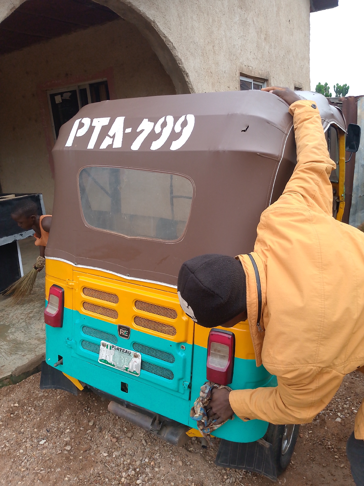 The image captures a scene in Jos, Nigeria, centered around a Bajaj RE tricycle, locally known as a "Keke." The Keke has a brown roof with the white lettering "PTA-799," a yellow middle section, and a turquoise lower portion. The license plate reads "PLATEAU" at the top. The last 4 digits of the license plate are 139.
A man in a black beanie and a mustard-yellow jacket is cleaning the right rear tail light of the Keke with a cloth. Another person, possibly a child, is visible on the left side of the Keke holding a broom and sweeping.
The background features a building with an arched entryway and windows. The ground is a mix of gravel and dirt.