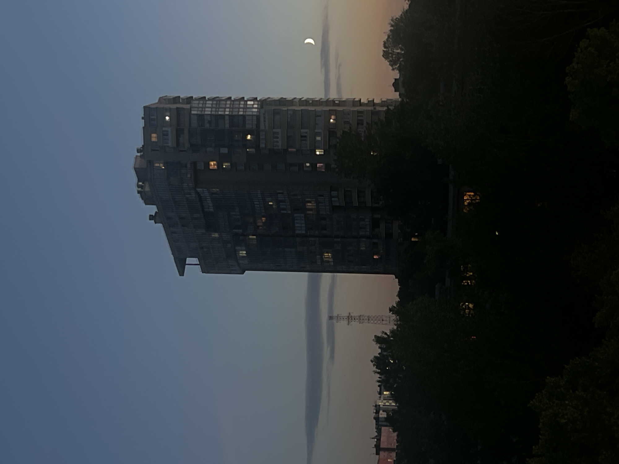 A tall, modern apartment building stands prominently in the center of the frame, its glass facade reflecting the twilight sky. Lights are visible in many of the windows, suggesting occupancy. To the left of the building, a cell tower rises above a line of trees, its structure silhouetted against the hazy sky. A crescent moon is visible in the upper right portion of the image, low on the horizon. The foreground is dominated by dark, leafy trees, obscuring any ground-level details and casting the scene in a twilight ambiance. The sky transitions from a deep blue at the top to a soft orange and pink near the horizon, indicating either dawn or dusk. The overall impression is one of a quiet evening in a residential urban area, possibly Sinyavino, Russia.
