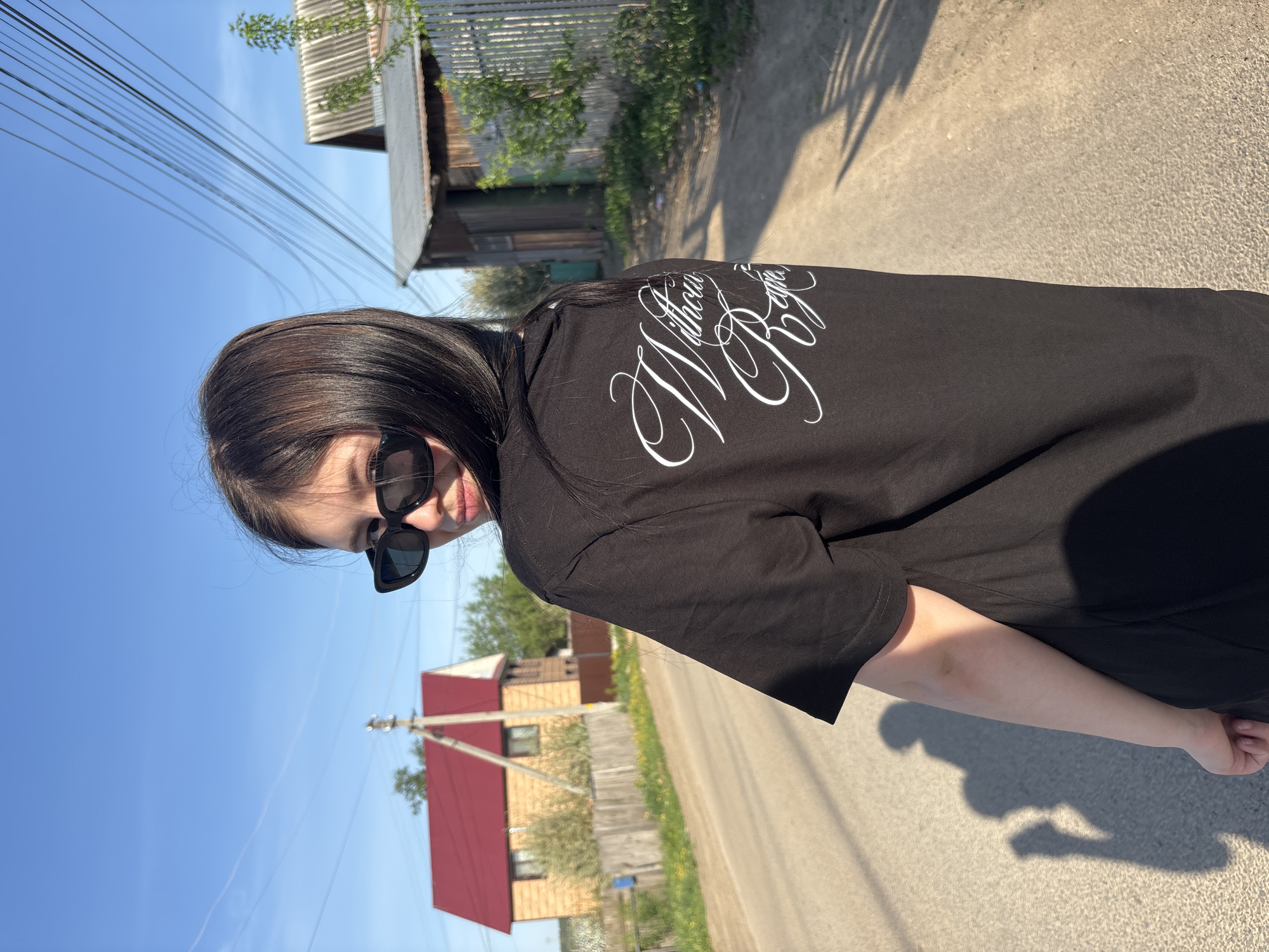 A young woman with dark hair and sunglasses is standing on a paved road in Фоки, Russia. She is wearing a black t-shirt with white lettering. Behind her are wooden houses and utility poles against a blue sky.
