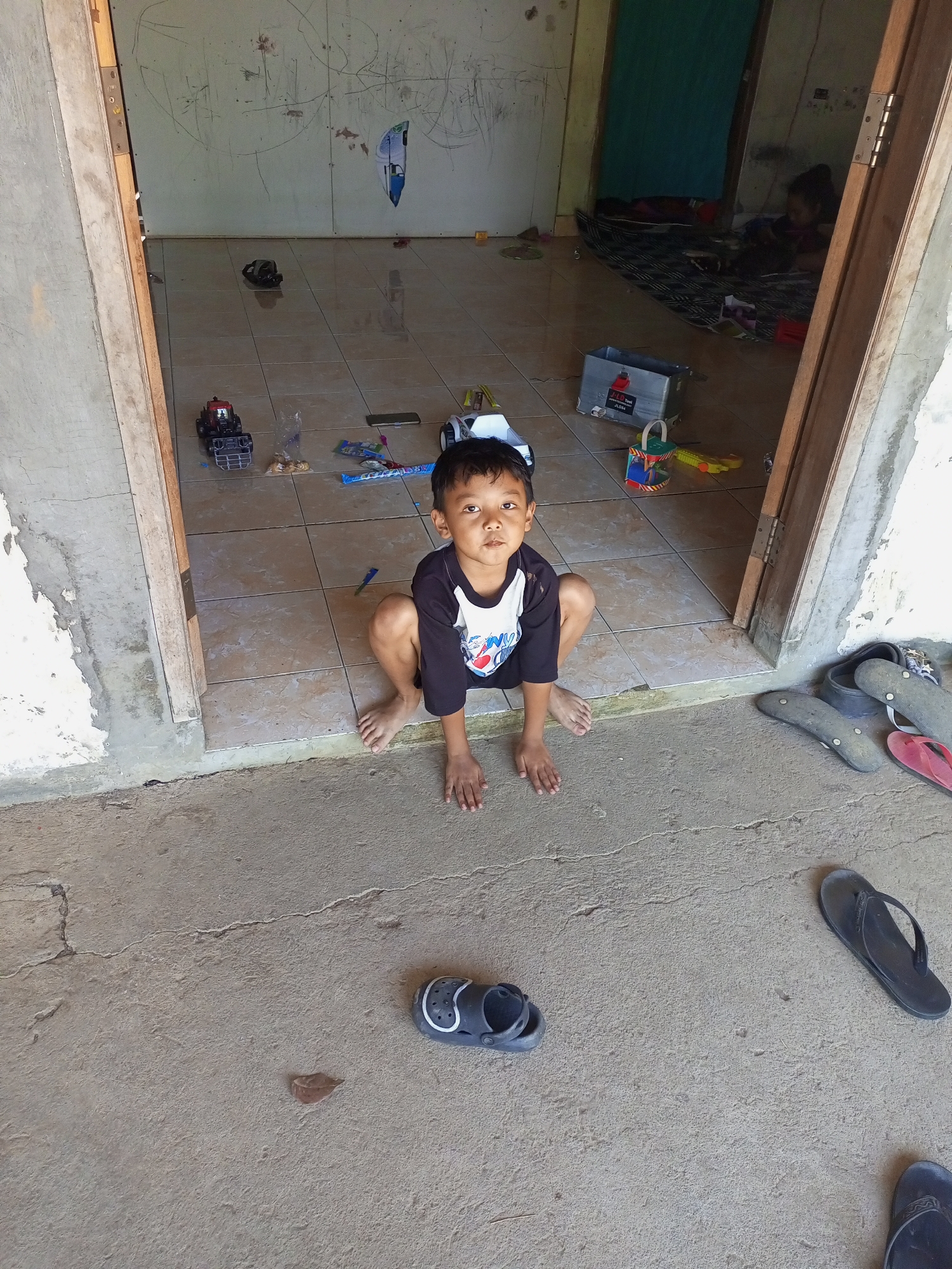 In a dimly lit room with a concrete floor, a young boy with short, dark hair and a black and white t-shirt kneels on the tiled entrance. He looks directly at the camera, his expression neutral. The room is sparsely furnished, with a mattress on the floor in the background and various toys scattered around, including a toy truck and a yellow toy. Several pairs of flip-flops and sandals are placed on the concrete floor outside the room. The walls are bare and appear to be made of concrete. The overall scene suggests a humble living environment in a rural area.