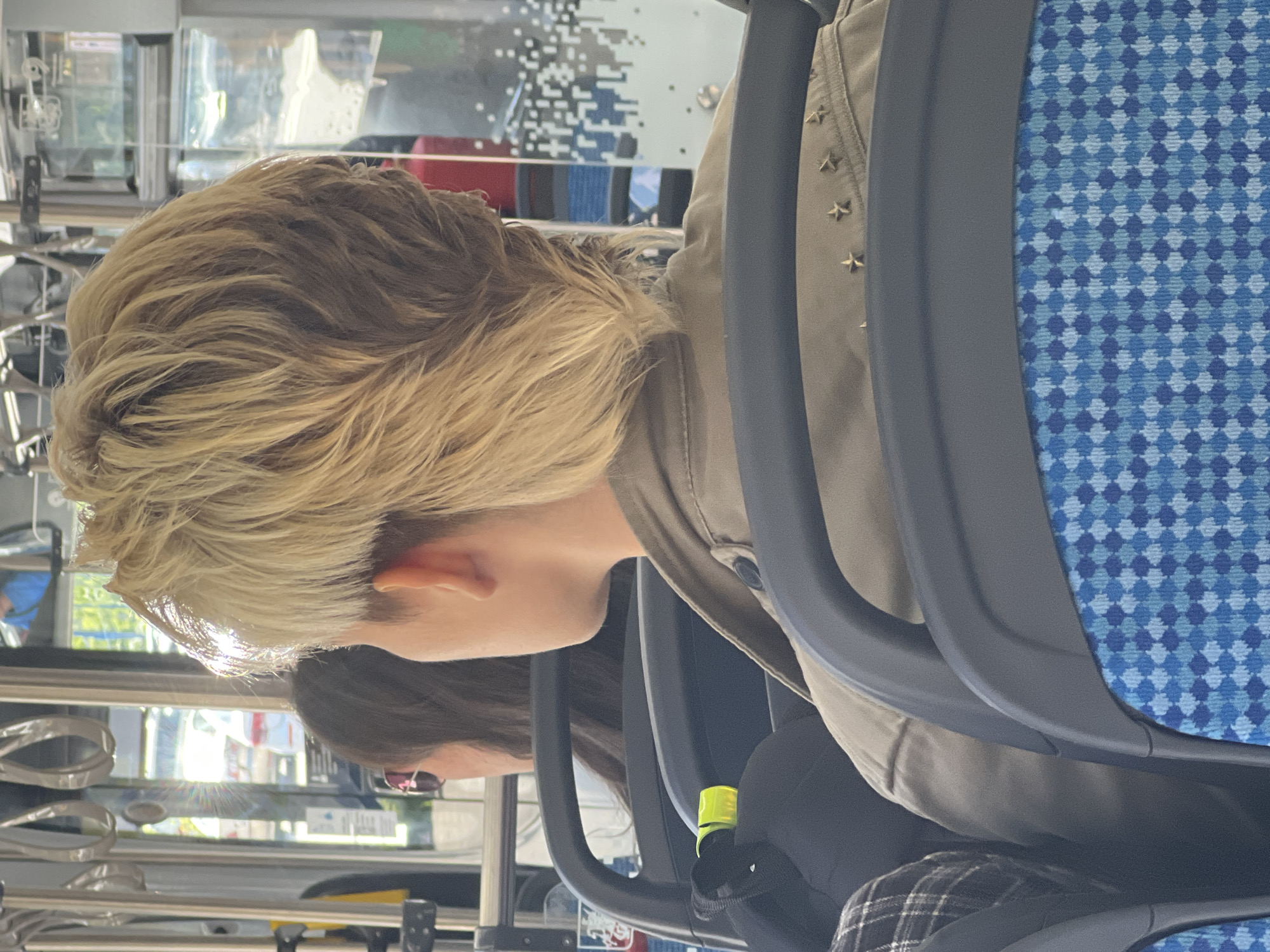 The image shows the back of a person's head with blonde, layered hair, wearing a khaki jacket with star embellishments. They are sitting on a bus in what appears to be Khimki, Russia, as indicated by the context. The bus interior is visible with blue patterned seats and handrails. Sunlight streams in from the windows, illuminating the hair. Another person with dark hair is visible in the background.