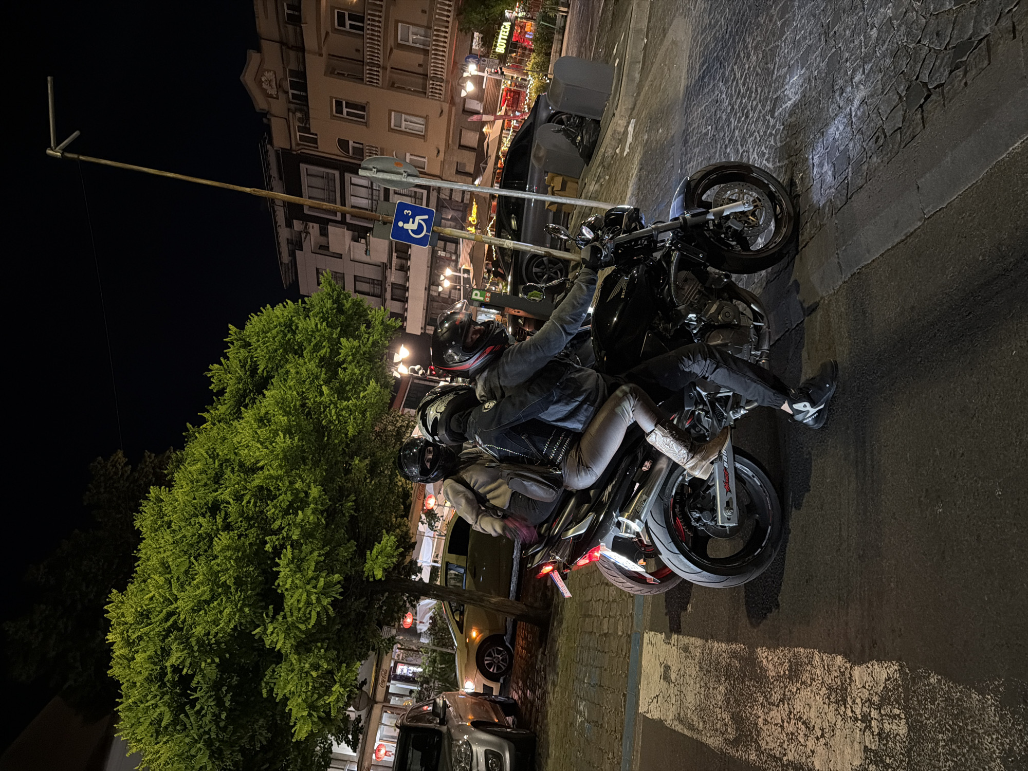 A group of three individuals are on a motorcycle at night on a street in Burgas, Bulgaria. The motorcycle is black and appears to be a sportbike. Two people are seated on the motorcycle, and one person is standing. The individuals are wearing helmets, and their faces are obscured. The background shows buildings, streetlights, trees, and other vehicles. There is a sign with a wheelchair symbol visible, indicating a disabled parking spot. The street is paved with cobblestones and asphalt. The lighting suggests it is nighttime or late evening.