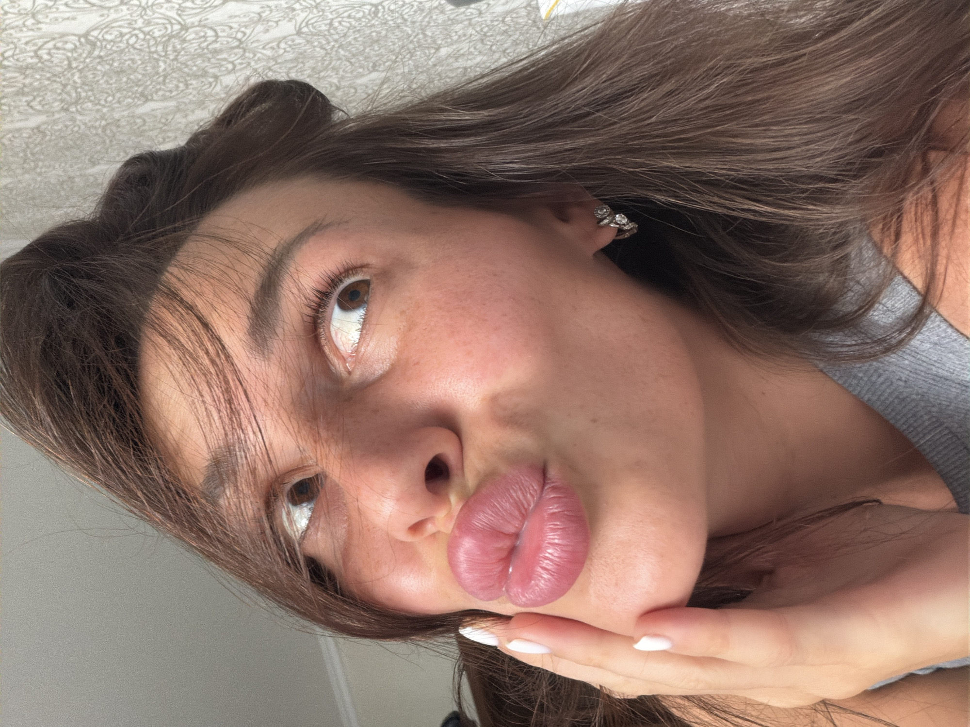 A close-up, indoor, selfie-style photograph shows a young woman with brown hair and brown eyes. She is looking upwards and to the left, with her lips pursed in a kissing gesture. Her right hand is placed on her cheek. She is wearing a grey tank top and a silver hoop earring with decorative stones in her left ear. The background is a patterned wallpaper. There is no visible indication of the location being Moscow, Russia.