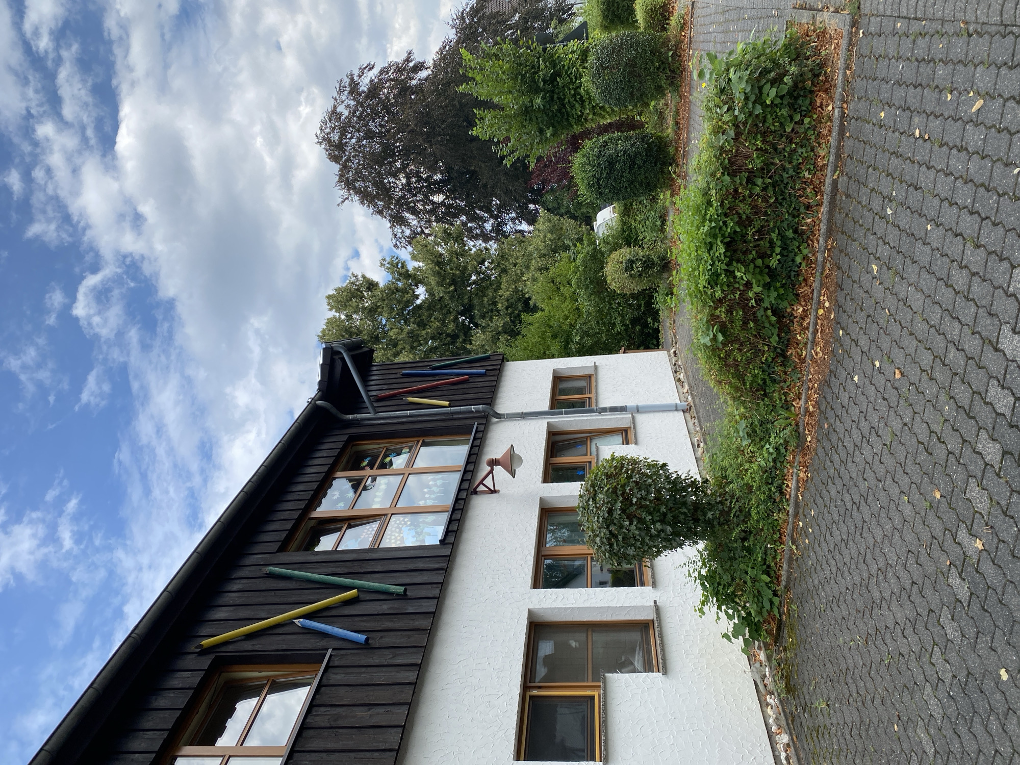 A close-up, low-angle exterior shot of a building in Flammersfeld, Germany, shows a white stucco facade with dark wood paneling on the upper level. Several windows with wooden frames are visible on both levels. Colorful cylindrical objects, resembling oversized pencils, are attached to the dark wood paneling. A single outdoor lamp with a reddish-brown fixture hangs from the wall below the upper windows. The building is adjacent to a gray paver driveway and a landscaped area with various bushes and trees. A large, dark-leafed tree dominates the background, with other green foliage and a cloudy sky completing the scene. No people or actions are depicted.