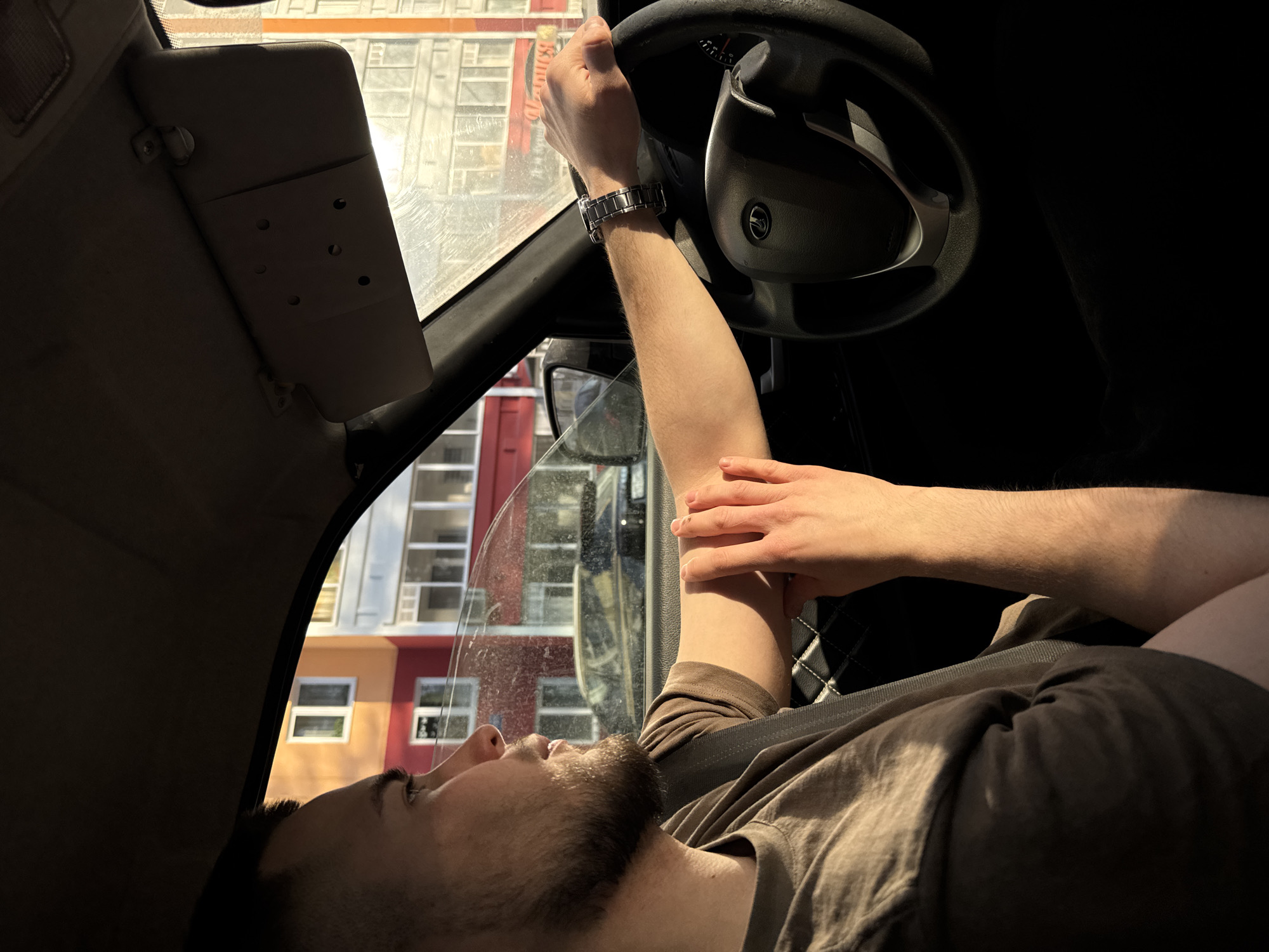 The image captures the interior of a vehicle from the driver's perspective, located in Perm, Russia. A young adult male with short dark hair and a beard is seated in the driver's seat, observed in profile looking forward and to the left. He is wearing a dark olive-brown short-sleeved t-shirt and a dark seatbelt across his chest. His left hand rests on his right forearm, which is positioned across his lap. His right hand firmly grips the black steering wheel, which bears a silver Lada logo. A silver-toned wrist watch is visible on his right wrist. The car's interior includes a beige sun visor in the upright position and a section of black quilted upholstery on the seat or door panel. Strong sunlight enters through the front and side windows, brightly illuminating the driver's right arm, the right side of his face, and the exterior environment, creating deep shadows within the vehicle. Through the car's windows, multi-story urban buildings are visible, featuring distinct orange and white, and red facades. Other vehicles are partially discernible in the background, suggesting a street or parking area.