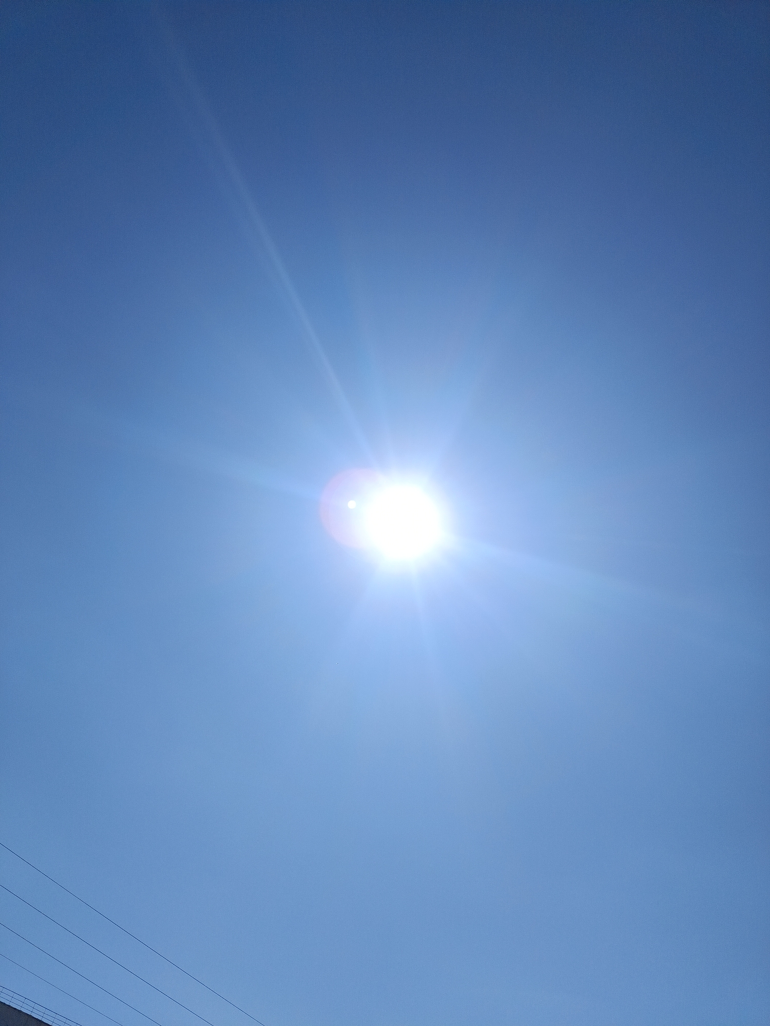 The image shows a bright sun centered in a cloudless, light blue sky. At the very bottom edge of the image, a portion of a structure (possibly a building) and power lines are visible. There are no people visible in the image. The exact location in Chaikovsky, Russia, cannot be determined with only the given context.