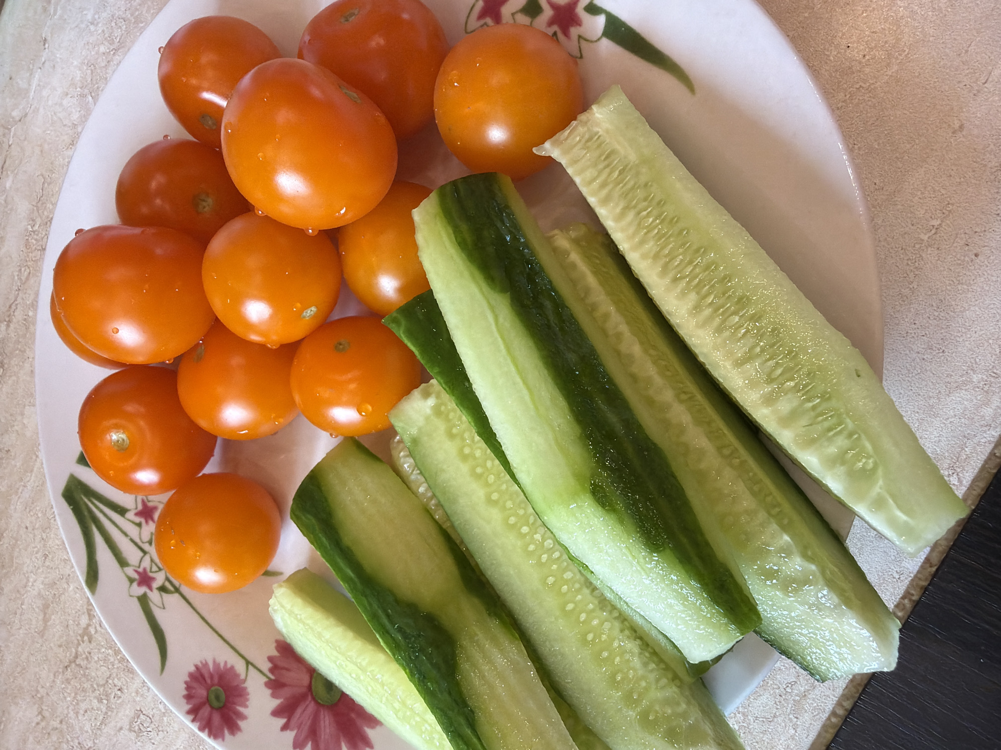 A close-up shot shows a white plate with a floral design filled with chopped cucumbers and cherry tomatoes. The cucumbers are cut lengthwise into long strips, revealing their white, seeded interiors and green skins. The cherry tomatoes are a vibrant orange-red and appear to be whole. The plate rests on a speckled beige countertop, with a dark wooden surface visible in the lower left corner. The lighting is bright, casting soft shadows. The scene is likely a kitchen or dining area. The image does not provide any information about the city of Solikamsk, Russia, or any specific actions or interactions.