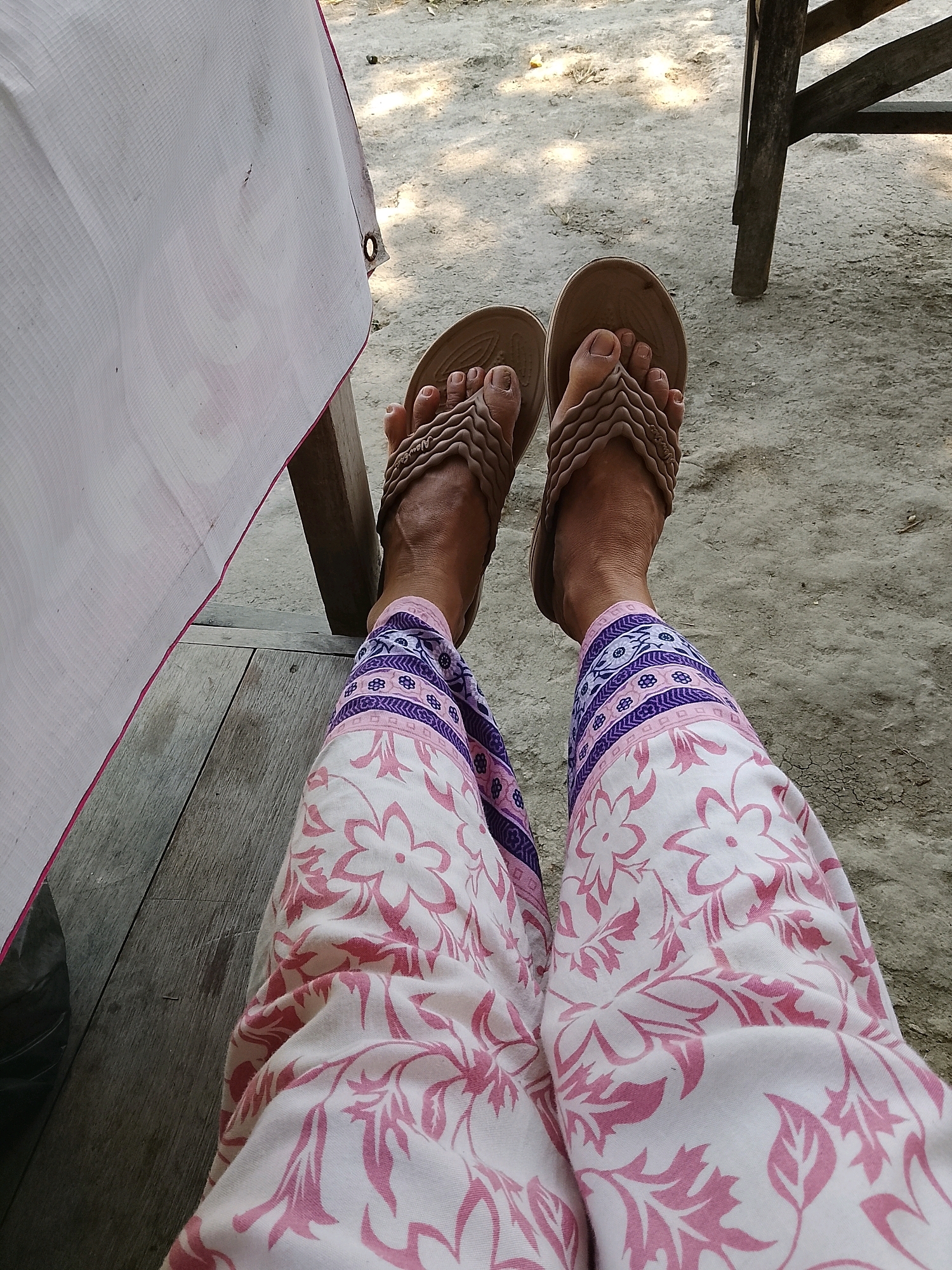 In Banaran, Indonesia, a person's legs are visible from the knees down, resting on a wooden surface. They are wearing patterned pants with a white background and pink floral designs, bordered with purple and blue geometric patterns around the calves. On their feet are brown flip-flops with a textured strap. The ground appears to be dirt, with sparse vegetation and scattered shadows from sunlight. To the left, a white fabric with red trim is draped over a wooden structure. To the right, the leg of a wooden chair is visible. The overall scene suggests a casual outdoor setting.