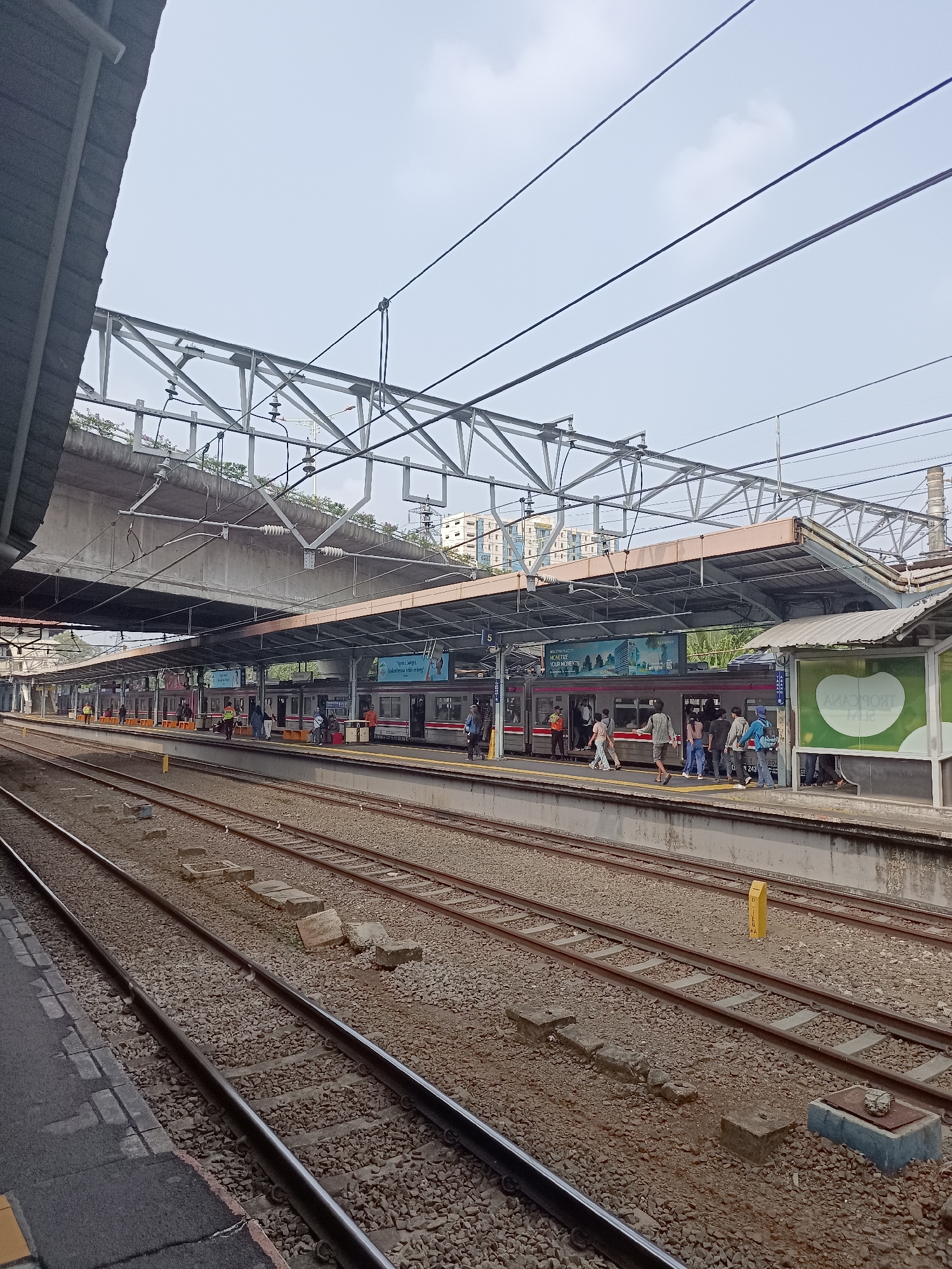 The image depicts a train station platform in Central Jakarta, Indonesia, under an overcast sky. Multiple train tracks run parallel to a covered platform where several people are visible. A train with red and white livery is stopped at the platform, with passengers boarding and disembarking. Some individuals are standing and walking, while others are seated. Advertisements are displayed on the side of the train and on the platform's overhead structure. Overhead power lines and a complex network of metal supports for the station roof are prominent. The platform itself is constructed with concrete and gravel ballast covers the ground around the tracks. Green signage is visible on a shelter structure on the right side of the frame. The overall impression is of a busy public transportation hub.