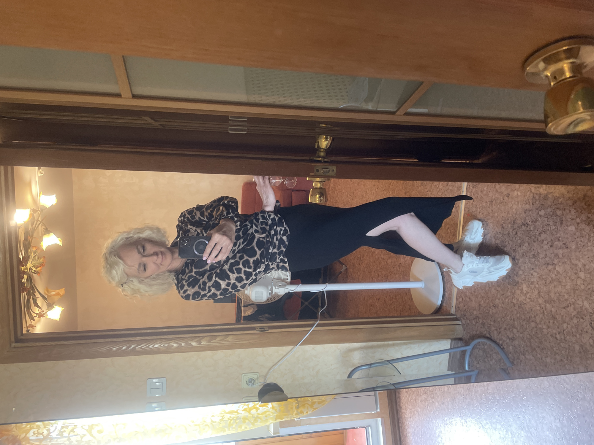 A woman with curly blonde hair is standing in front of a mirror in a room. She is wearing a leopard-print top and a black skirt with a high slit. She is also wearing white sneakers. She is holding a phone in her right hand and appears to be taking a selfie. To her left, a white fan is visible. The room has light-colored walls and a wooden door frame. There is a light fixture with orange lampshades on the ceiling. The floor is covered in a speckled brown material. The image is taken indoors and there is no visible indication of the location being in Saint Petersburg, Russia.